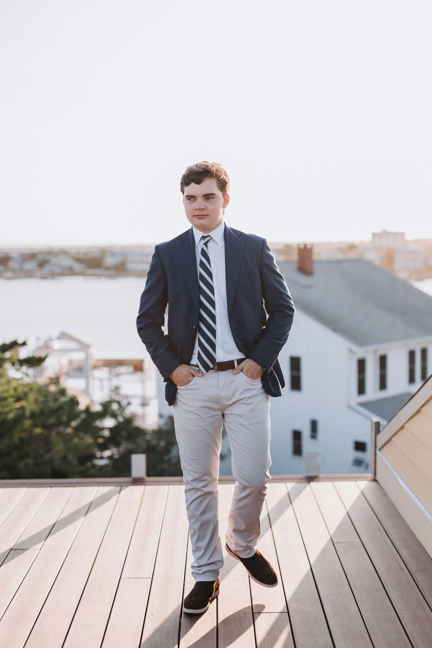Young man in navy blazer and striped tie standing on wooden deck overlooking coastal homes.