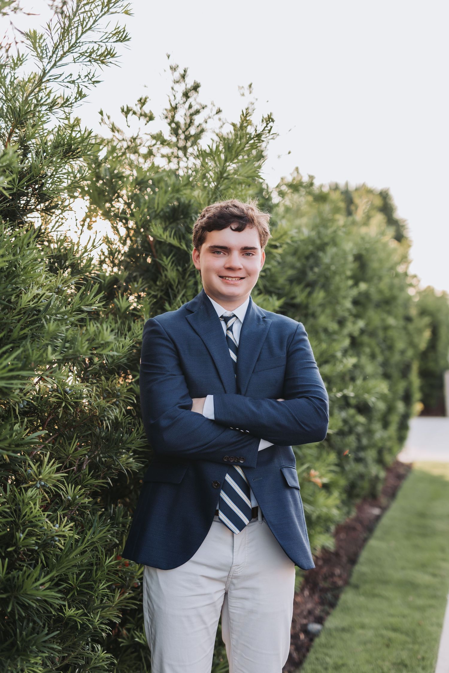 Formal portrait of someone in navy blazer and tie standing next to landscaped greenery.