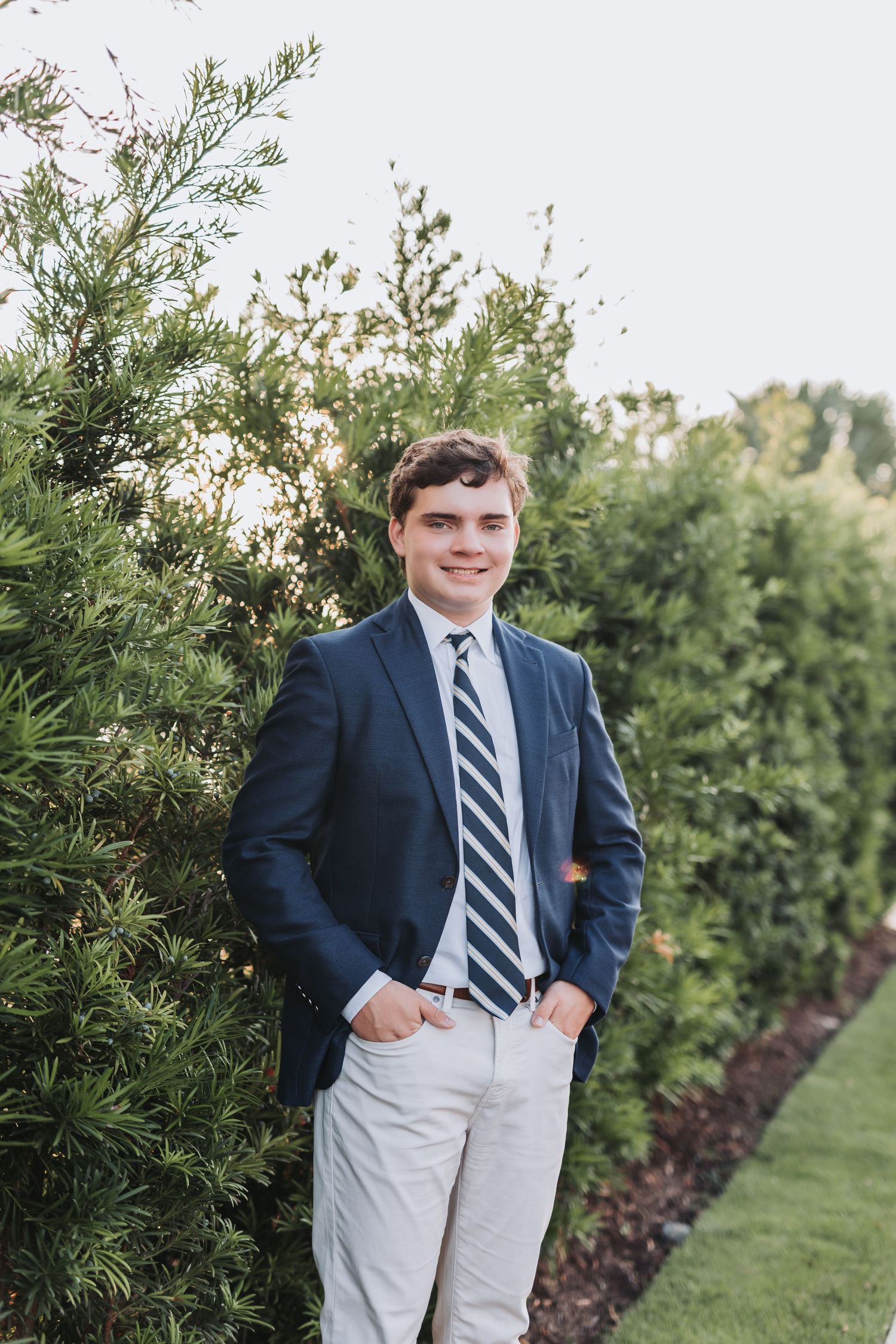 Professional photo of someone in navy suit and striped tie posed by landscaping bushes.