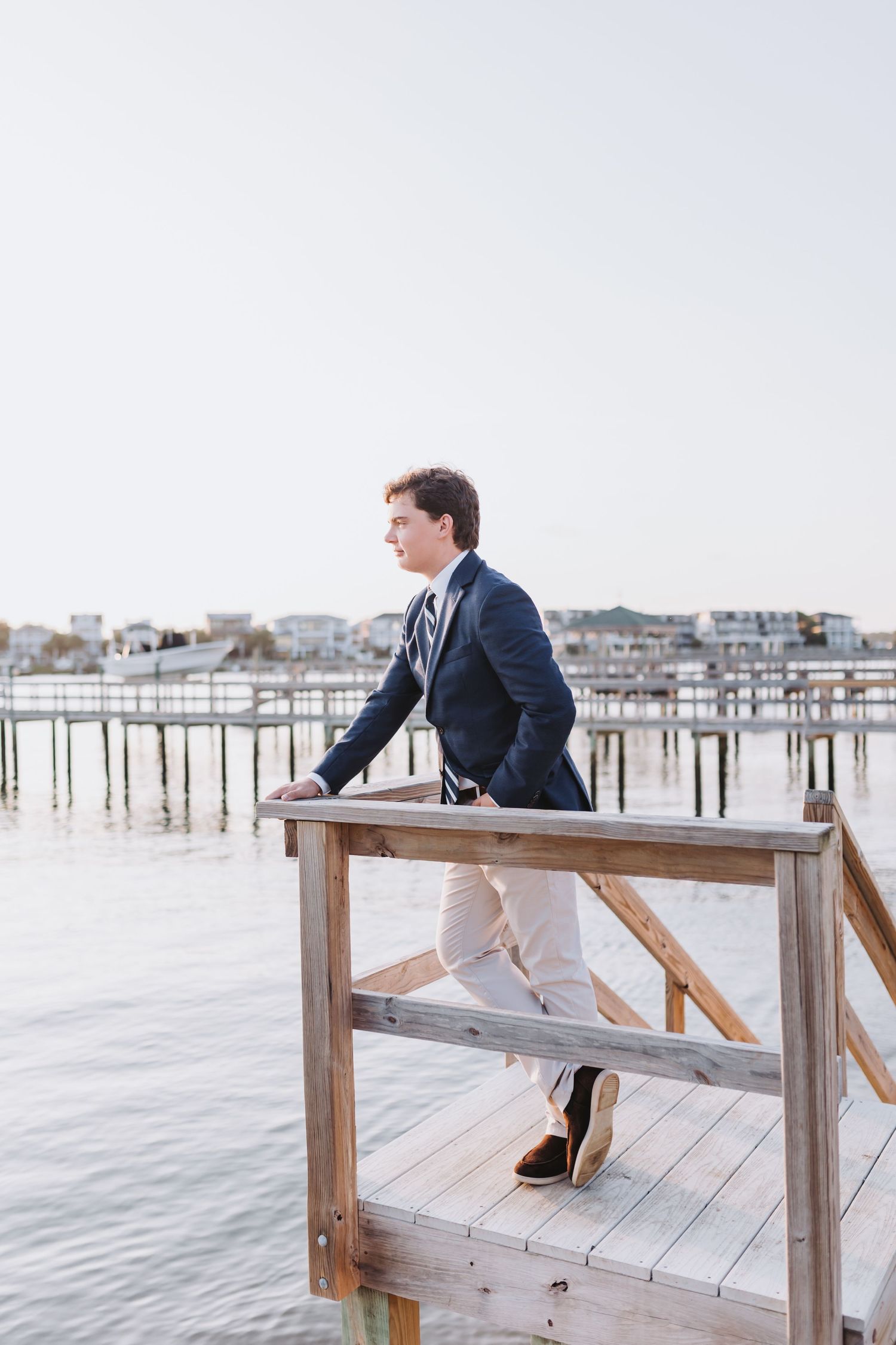 Person in formal attire leaning on wooden dock railing overlooking marina at sunset.