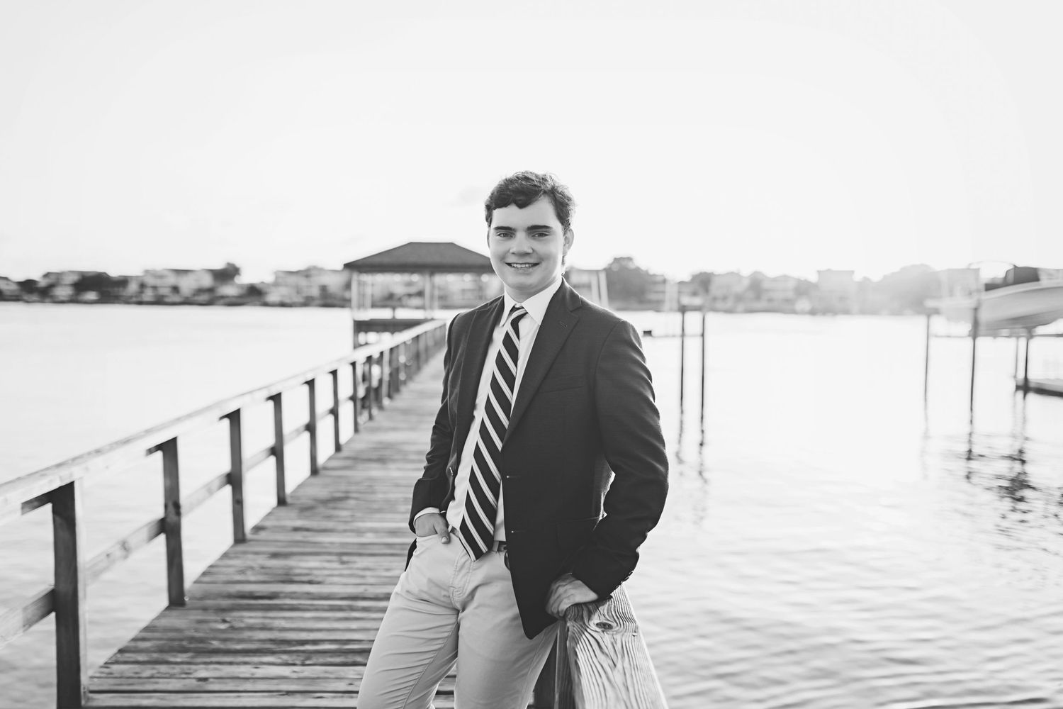 Portrait on a long wooden pier with water and coastal homes visible in the background.
