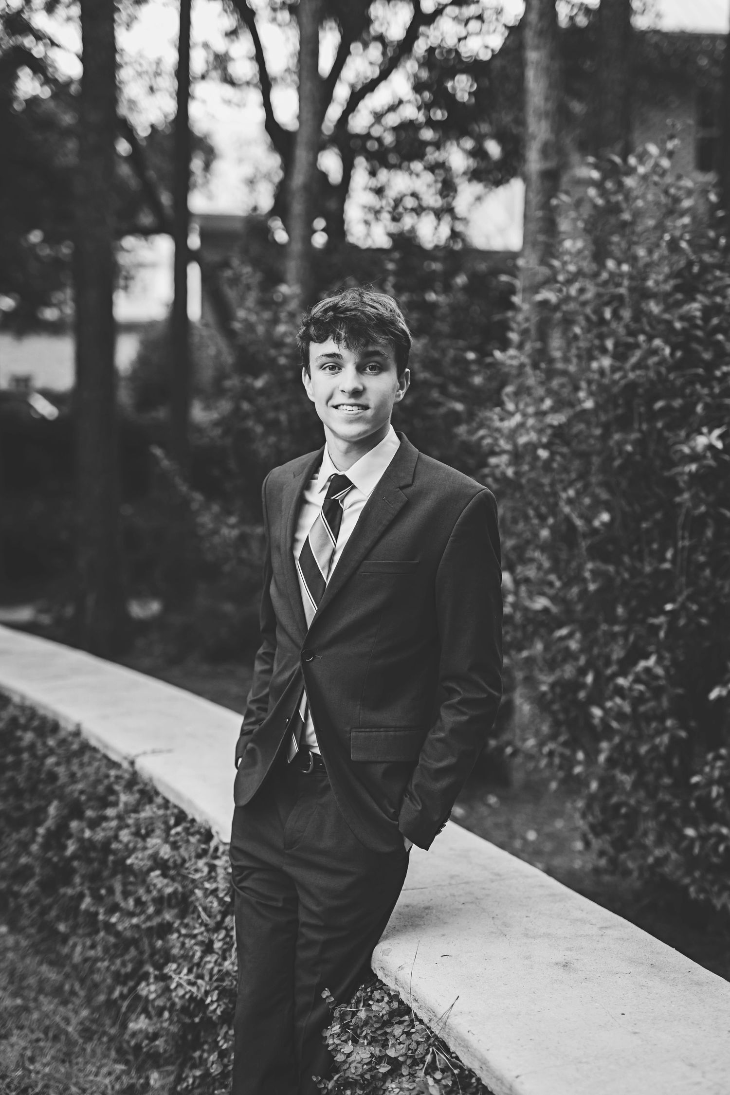 A person in a dark suit poses on a garden path surrounded by lush greenery.