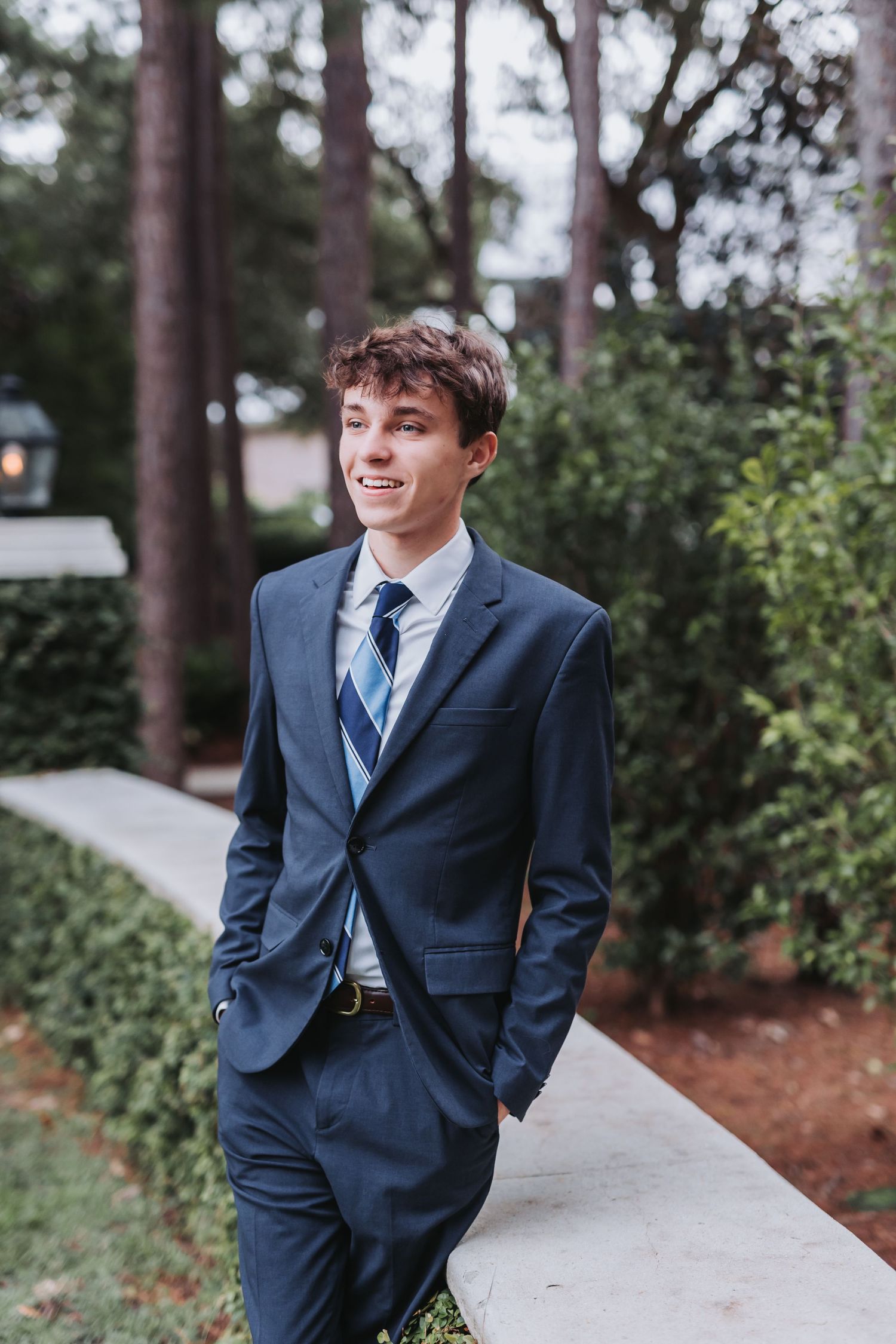 Professional portrait in a navy suit and tie on a landscaped pathway with pine trees.