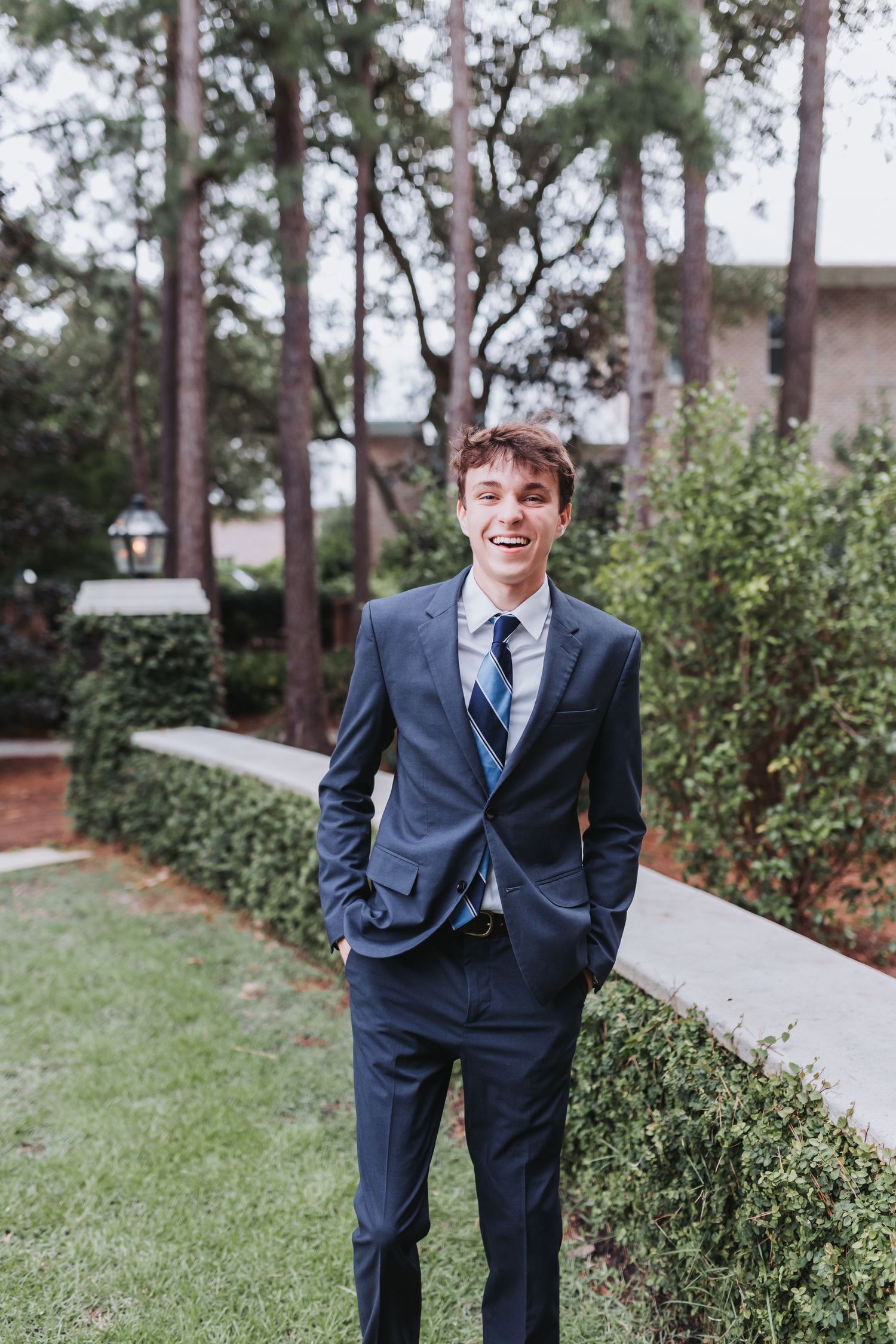 Formal portrait in a navy suit along a garden path with tall pine trees in the background.