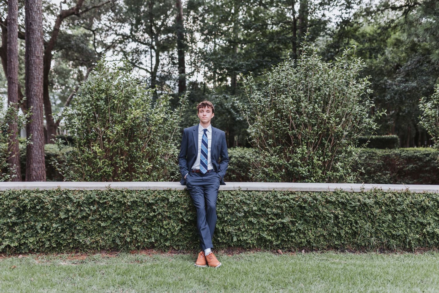 Portrait of someone in a blue suit leaning against a hedge wall with pine trees in background.