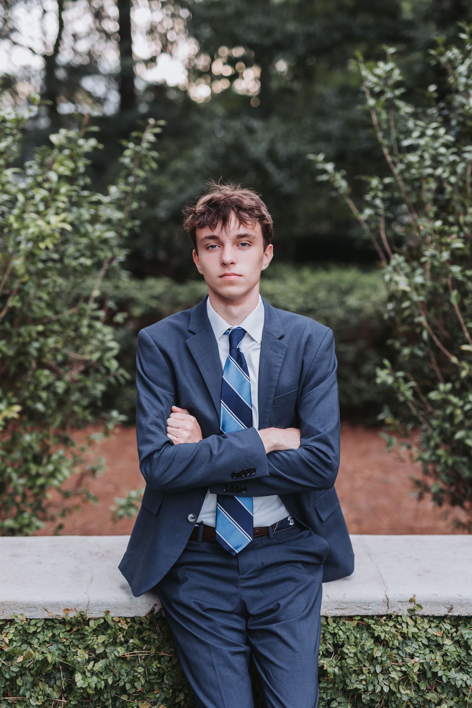 Young person in navy suit and striped tie poses confidently against greenery background.