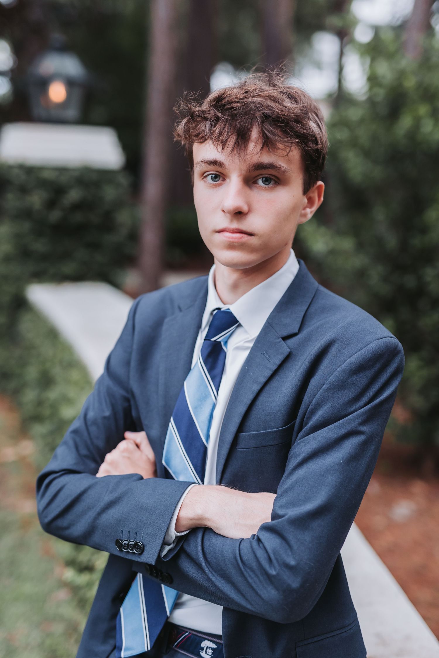 Professional portrait of person in blue suit with crossed arms against outdoor setting.