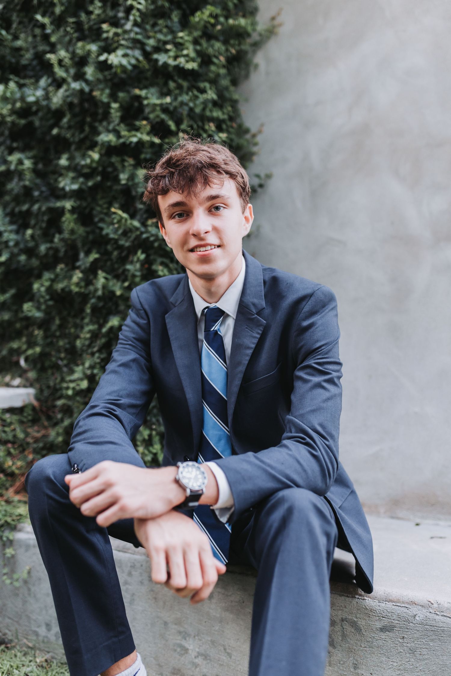 Casual seated portrait showing blue suit and striped tie against concrete wall.