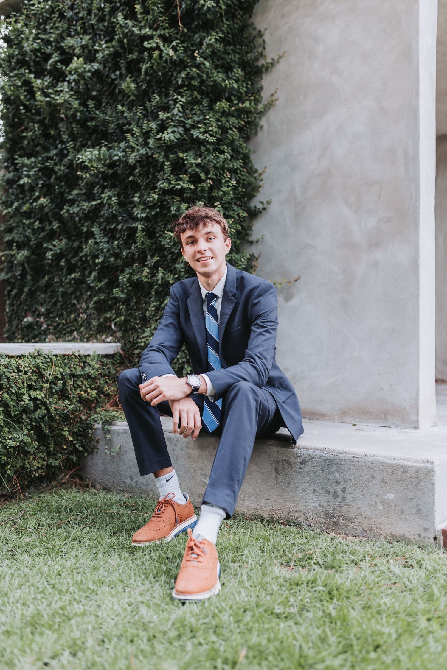 Casual seated pose showing brown shoes and navy suit against garden backdrop.