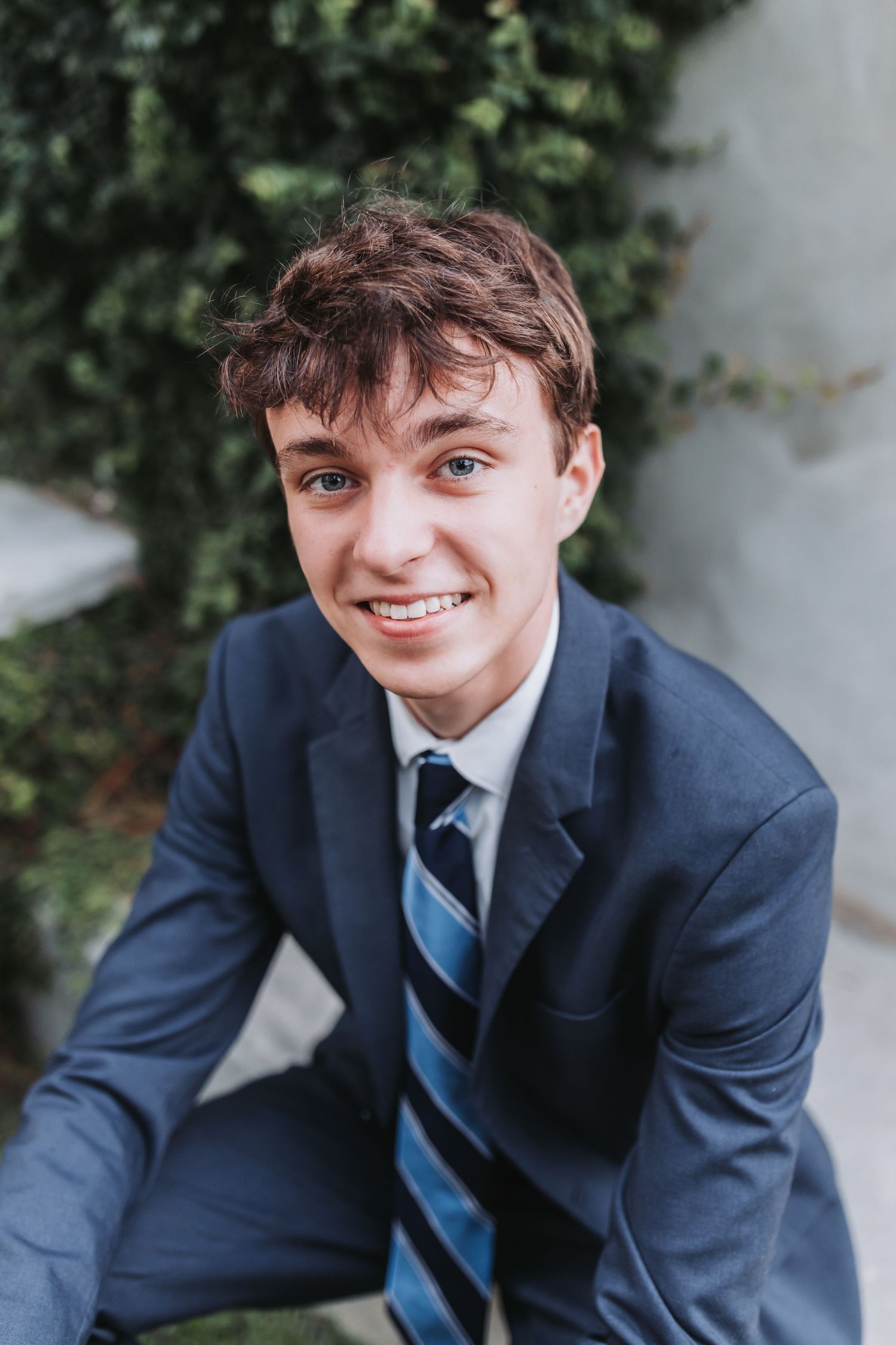 Close-up portrait showing bright expression and navy suit against garden background.