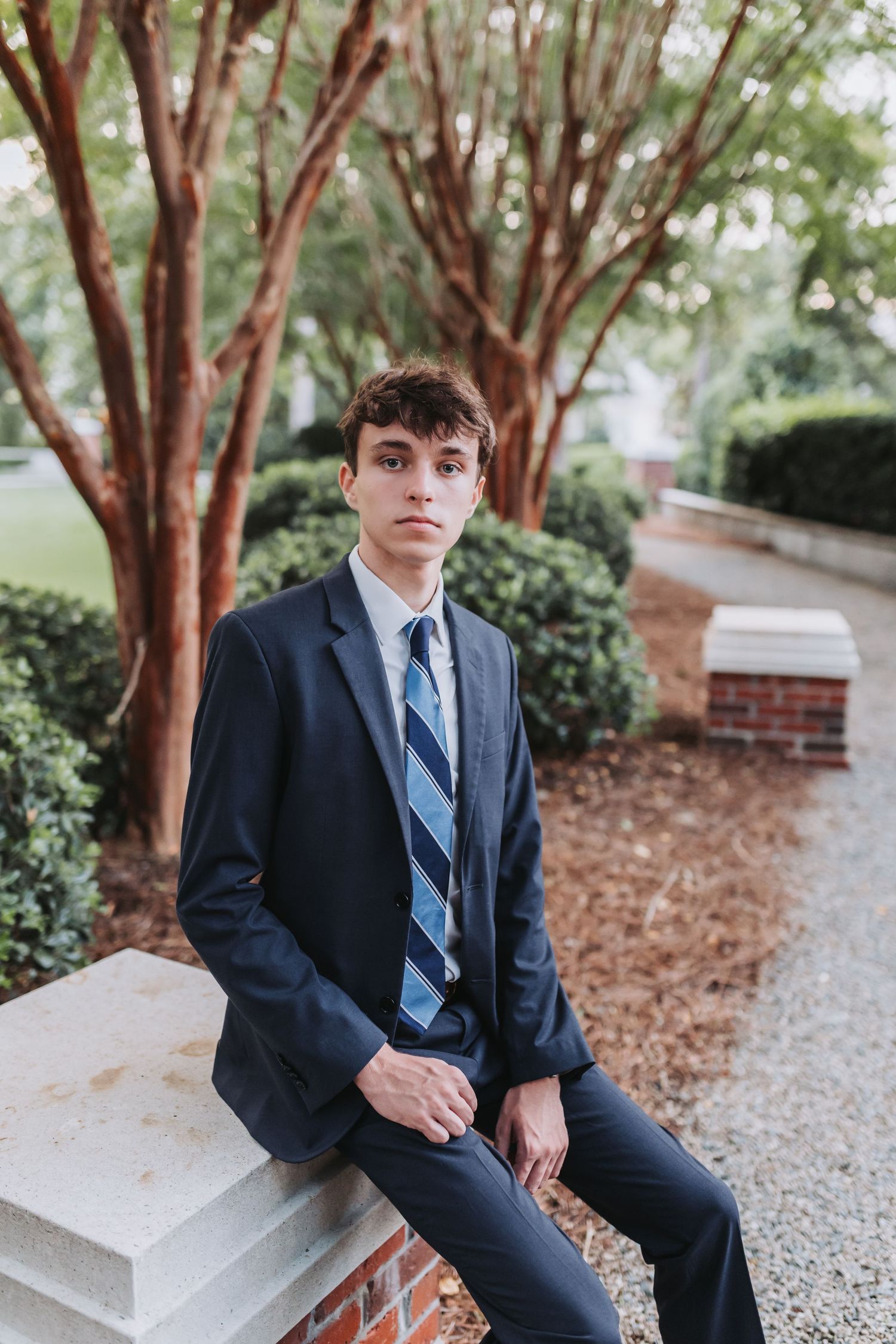 A young person in a navy suit sits casually on a brick ledge surrounded by trees in a formal garden setting.