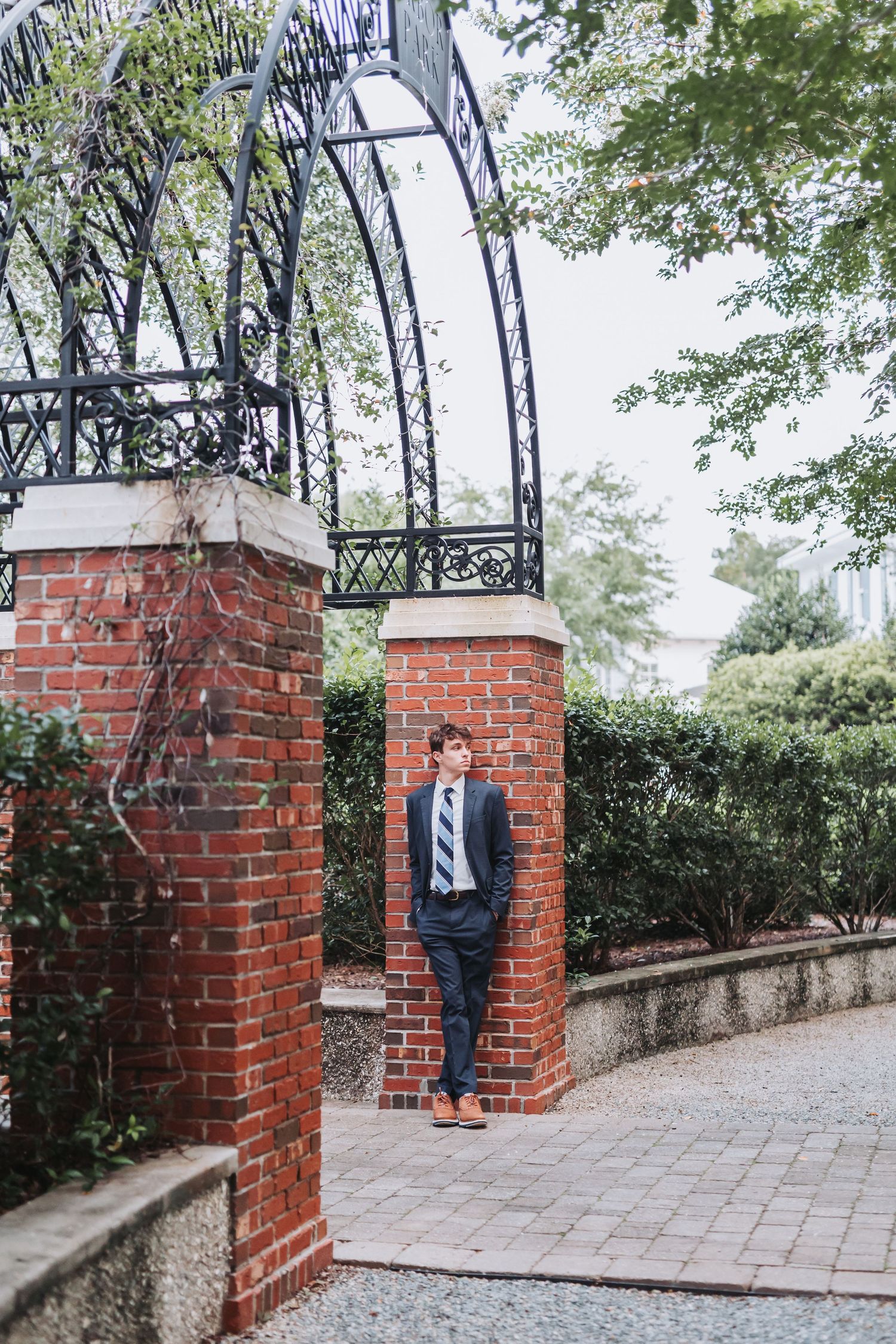 A figure in a dark suit stands between brick pillars beneath an elegant iron archway.