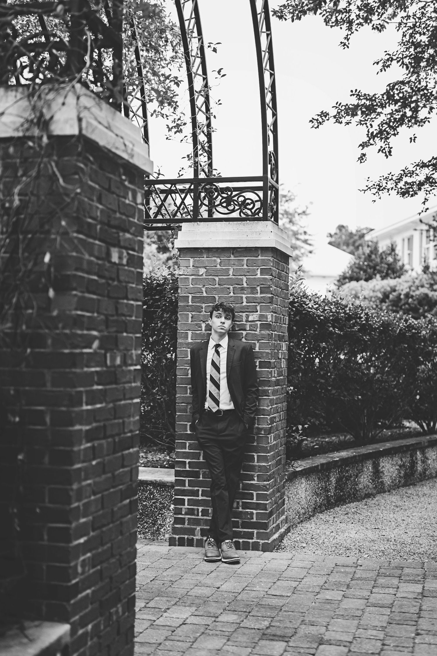 A figure in a tie stands between brick pillars on a paved garden path.