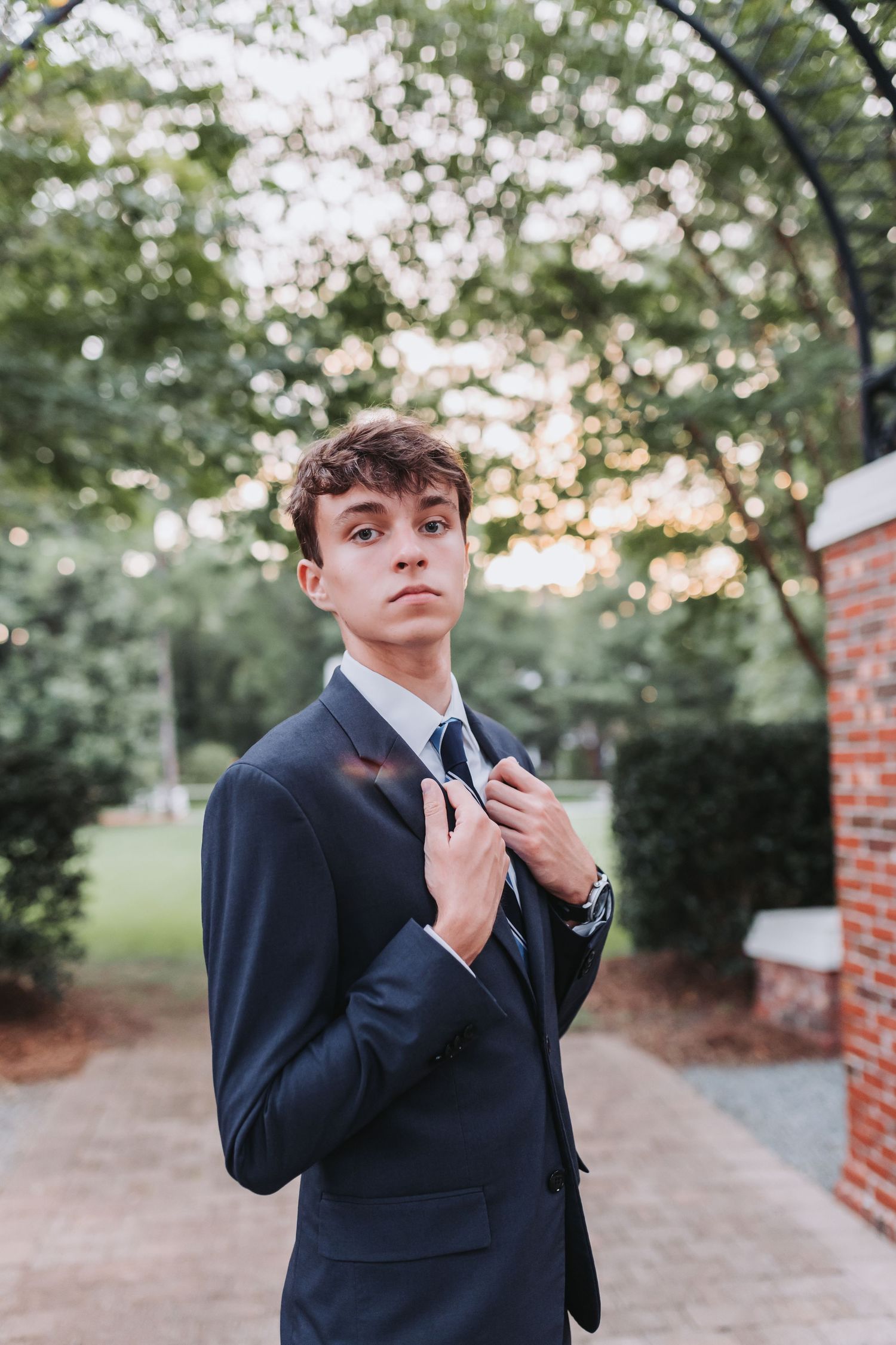 A portrait shot against blurred trees with warm sunset lighting and brick architectural elements.