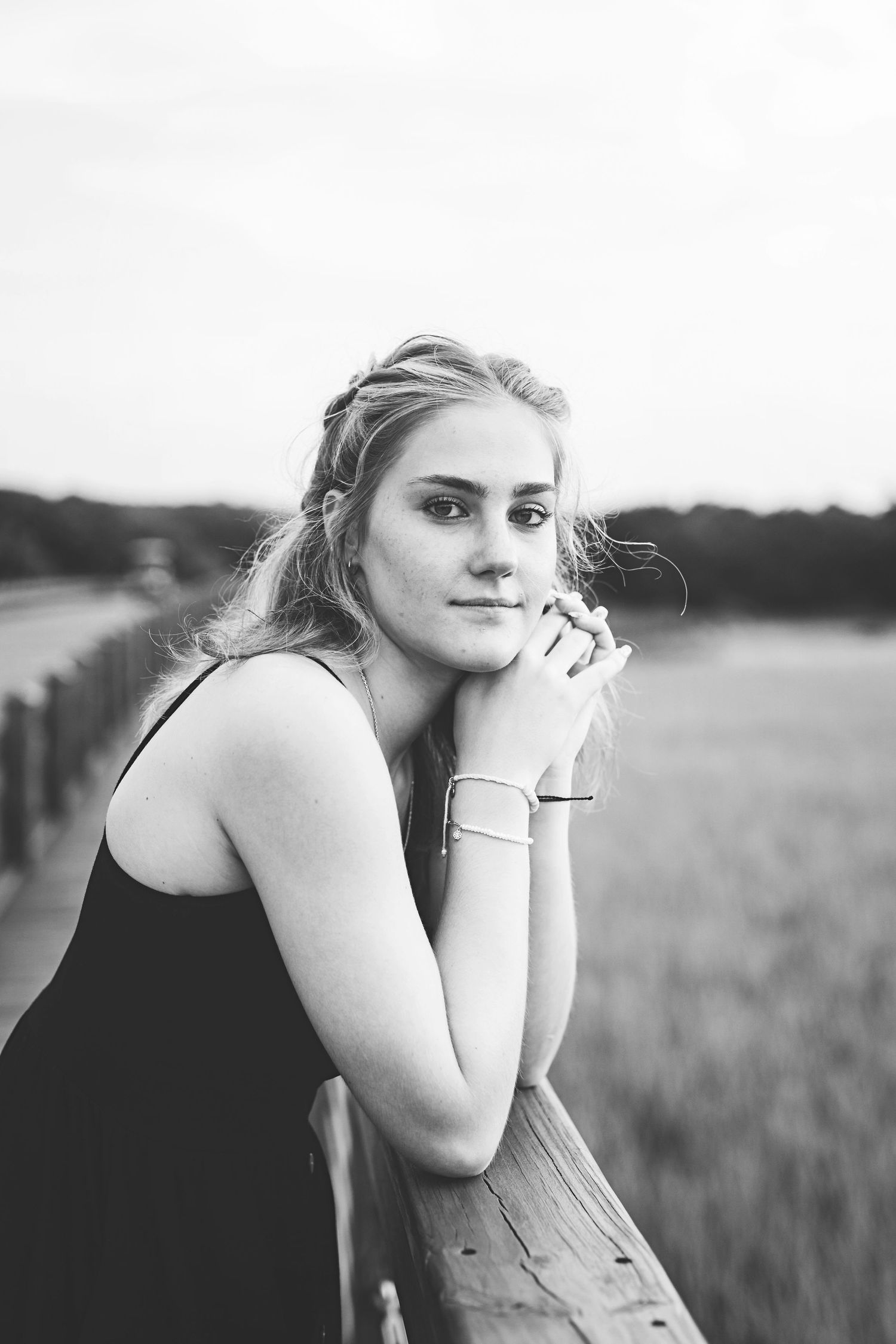 Black and white portrait of someone in black dress leaning on wooden railing at sunset.
