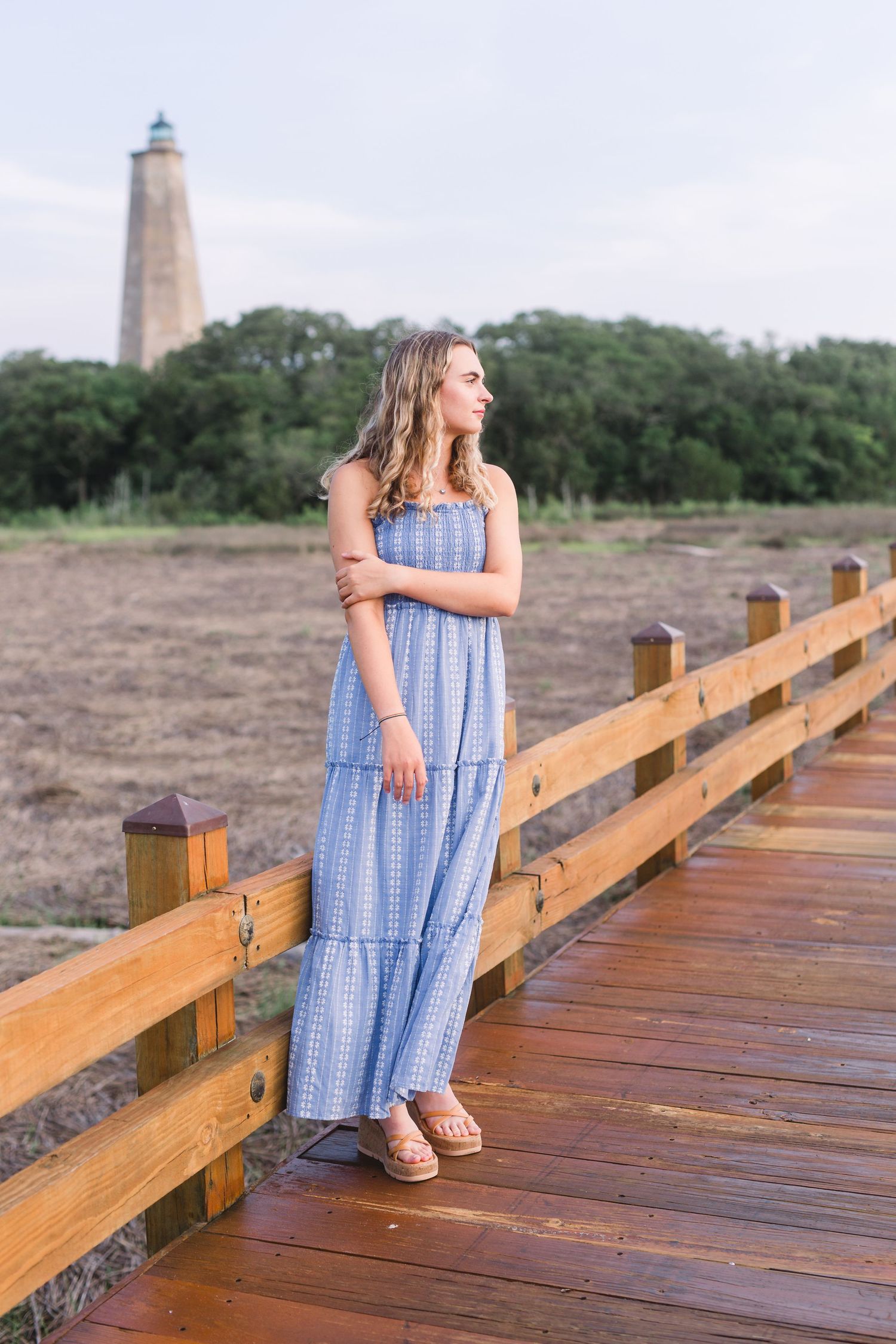 Blue maxi dress silhouette against lighthouse and wooden boardwalk at sunset.