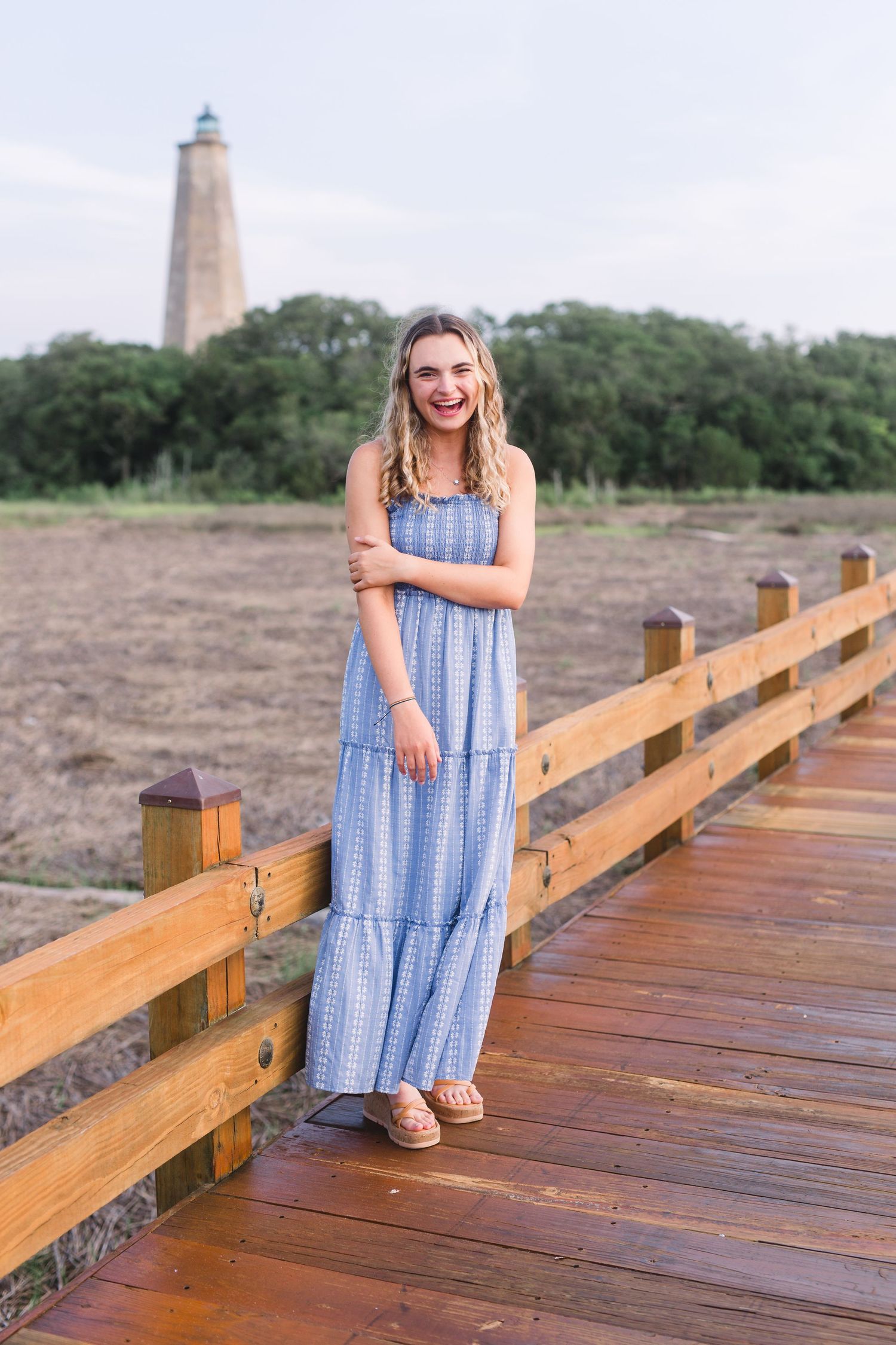 Person in flowing blue dress poses on wooden boardwalk with lighthouse in background.