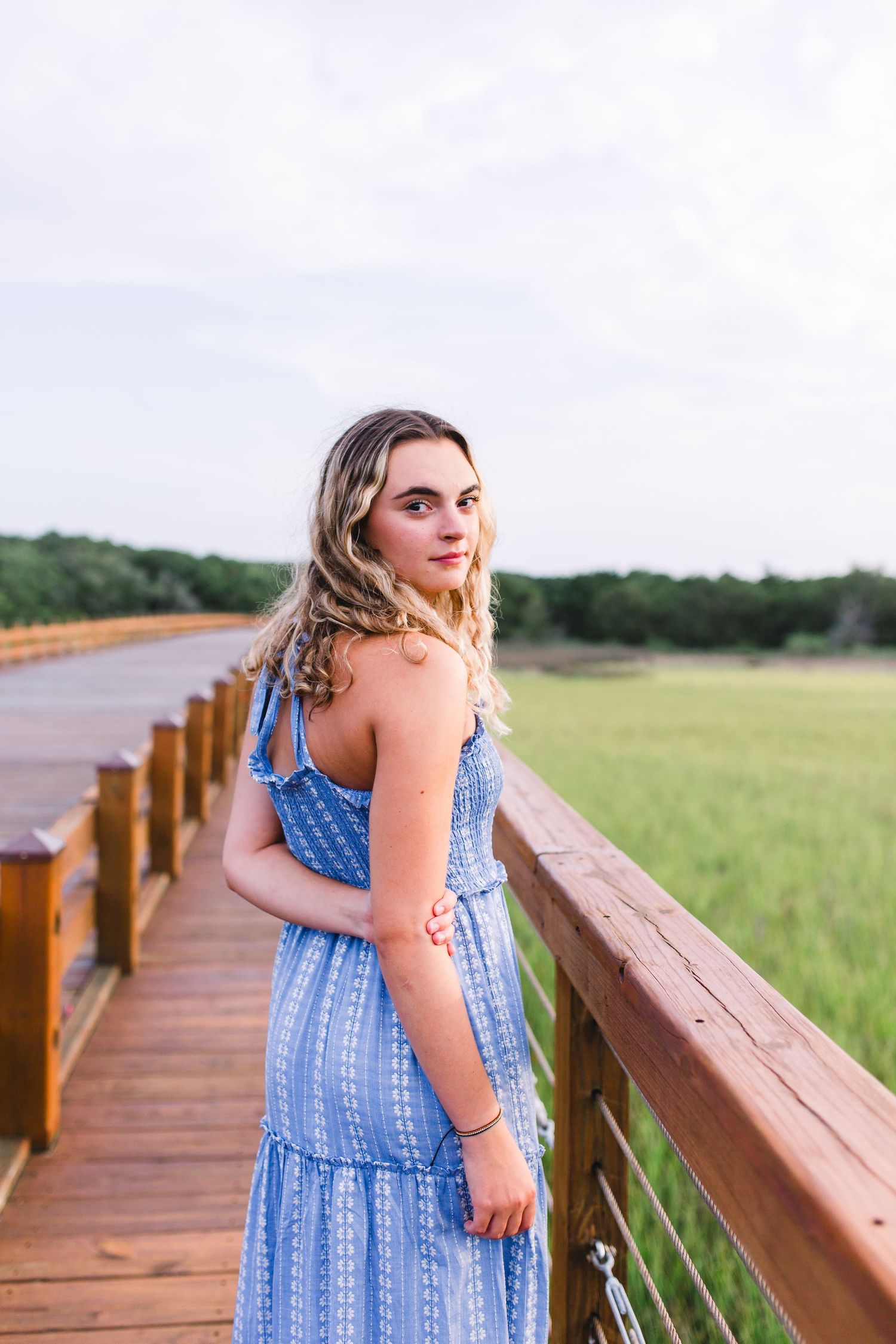 Casual pose in blue summer dress on wooden boardwalk overlooking coastal landscape.