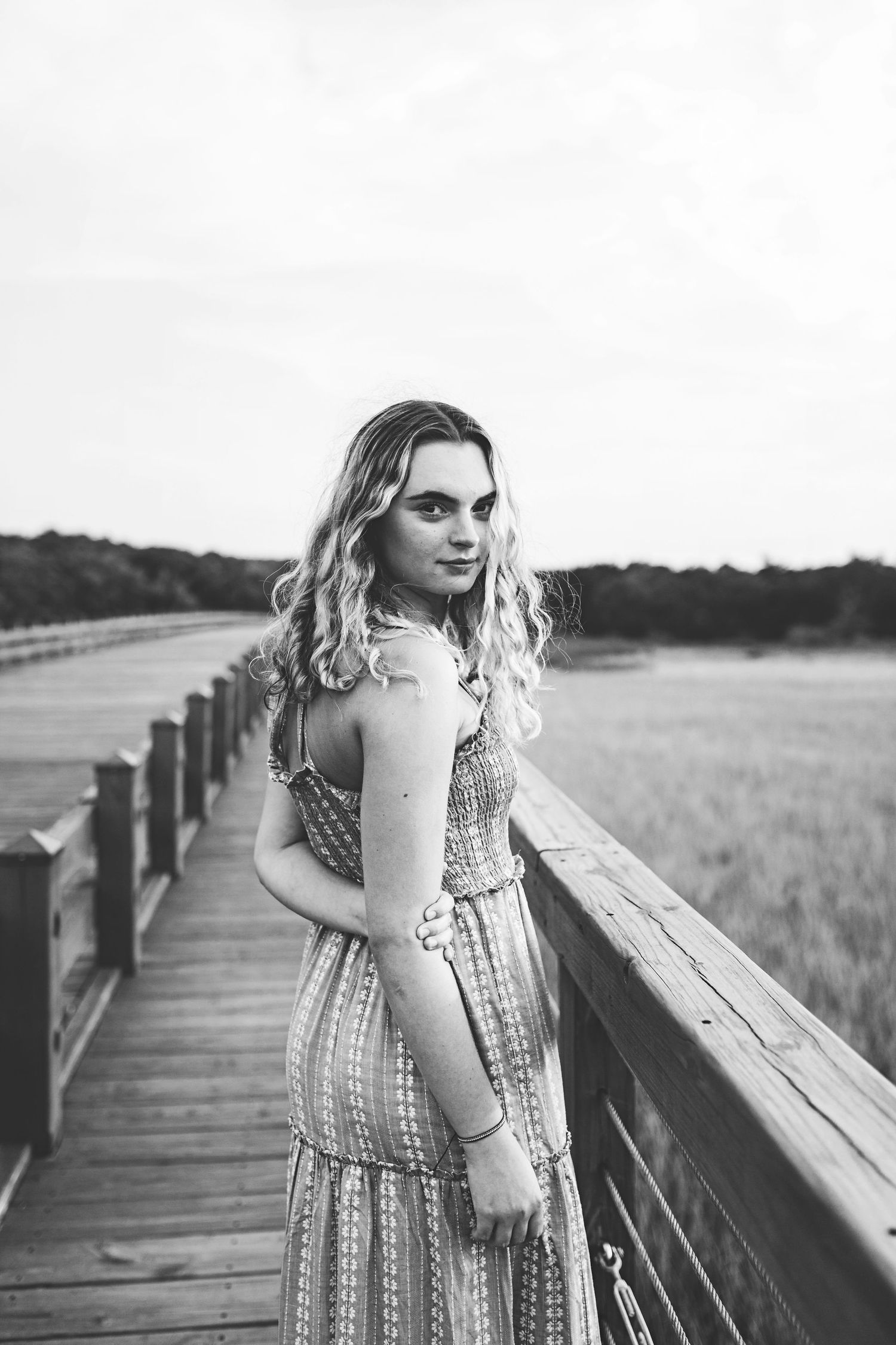A woman in a patterned dress poses on a wooden boardwalk overlooking a meadow in black and white.