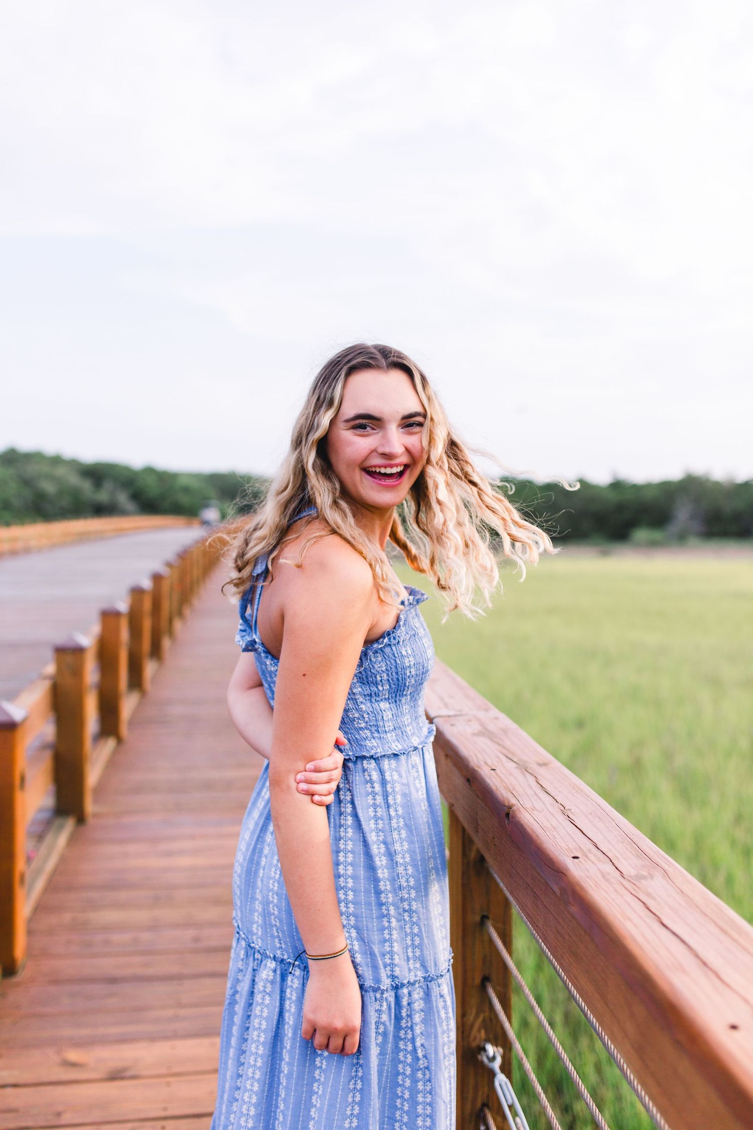 A woman in a blue and white patterned dress smiles while standing on a wooden boardwalk during golden hour.