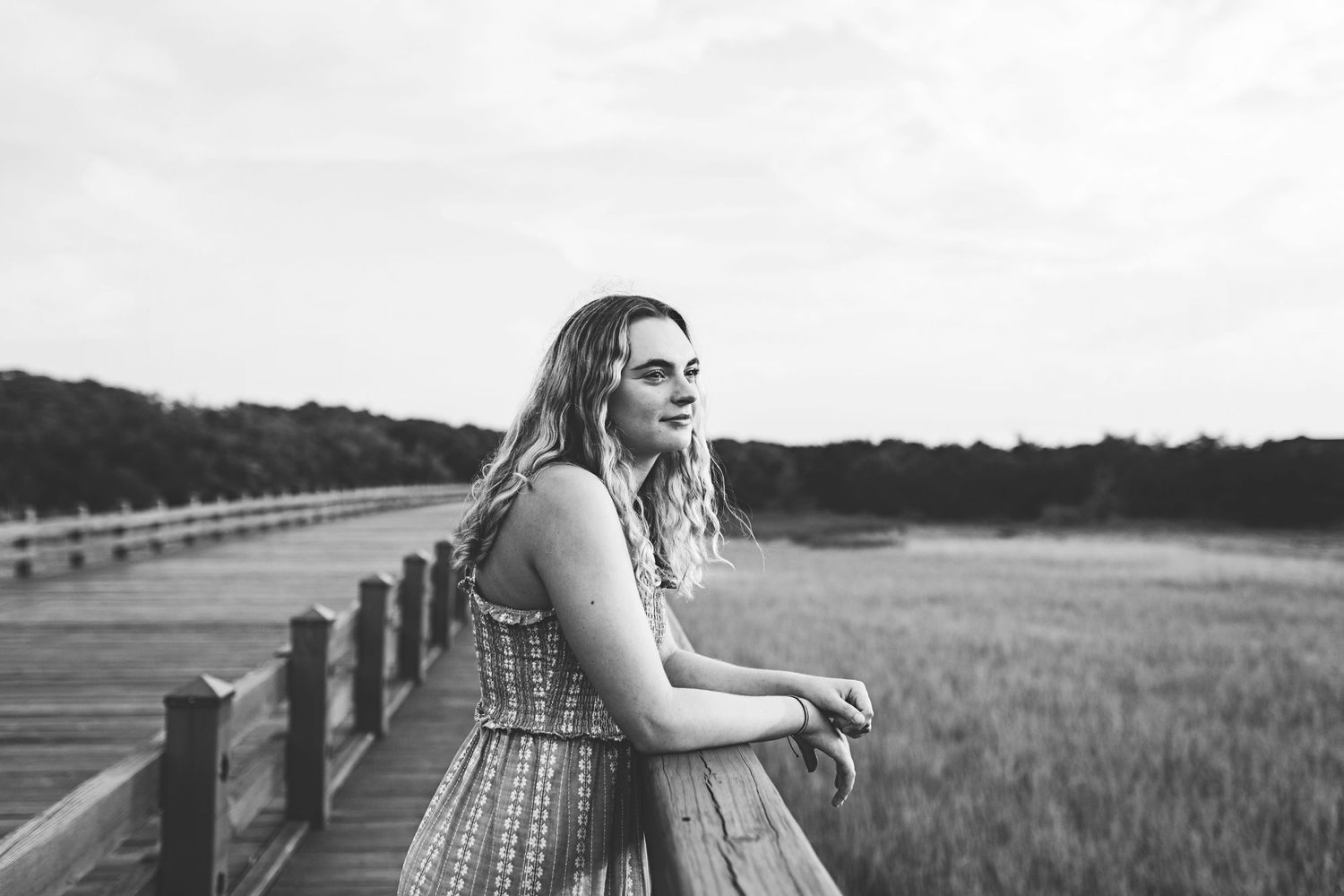A woman in a summer dress leans against a wooden railing overlooking a scenic marsh landscape.
