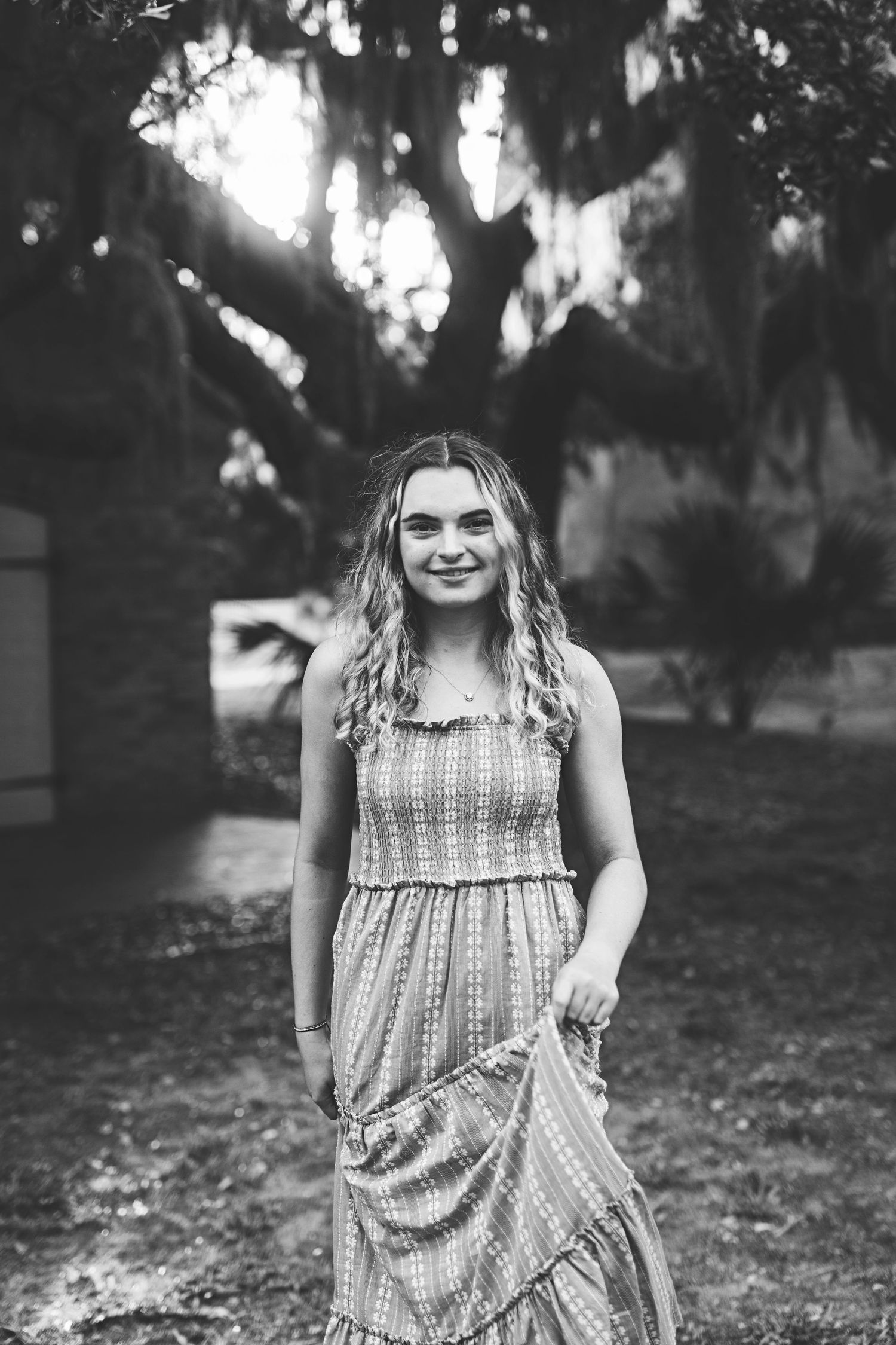 A black and white portrait of a woman in a patterned dress standing in a garden setting.