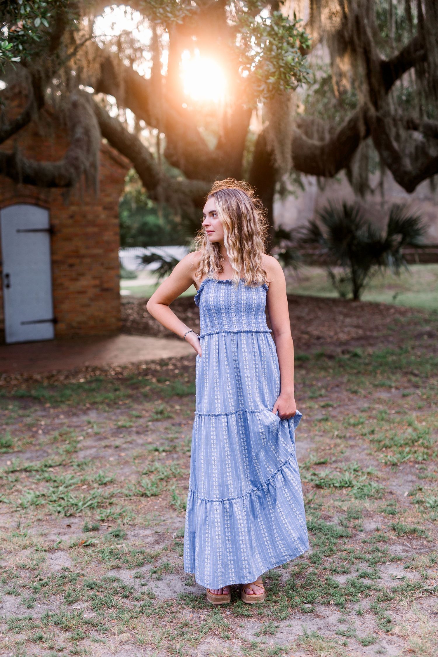 A woman in a tiered blue dress poses in a garden with Spanish moss and sunset lighting.
