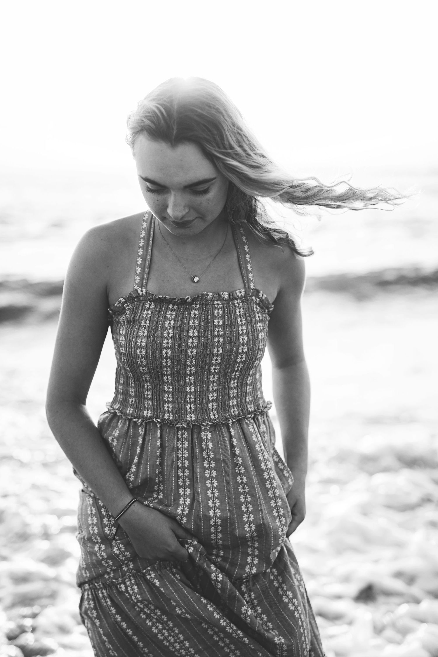 A black and white beach portrait of a woman in a patterned sundress looking down.