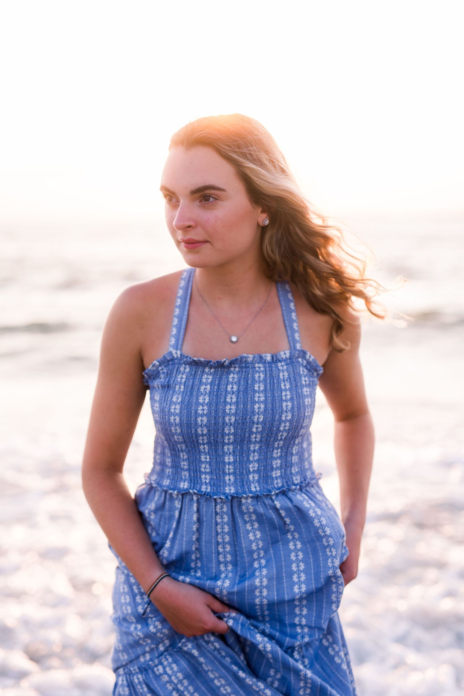 A woman in a blue patterned dress poses on the beach during golden hour.