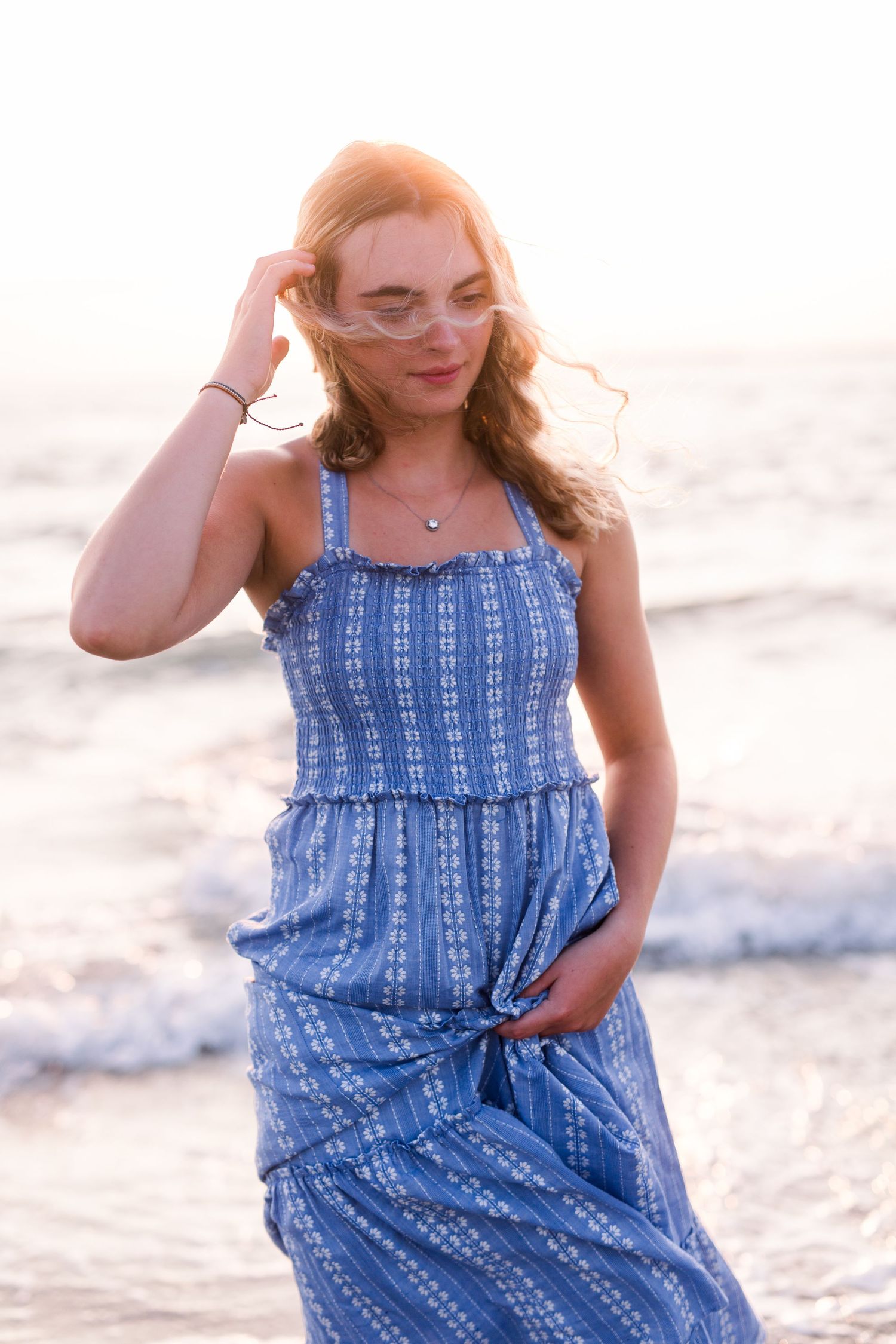 A woman in a blue summer dress smiles while touching her hair on a beach at sunset.