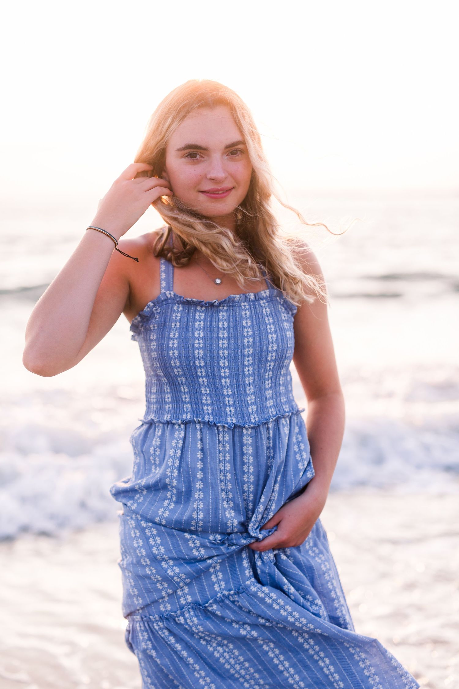 A woman in a blue striped sundress poses playfully on the beach at sunset.