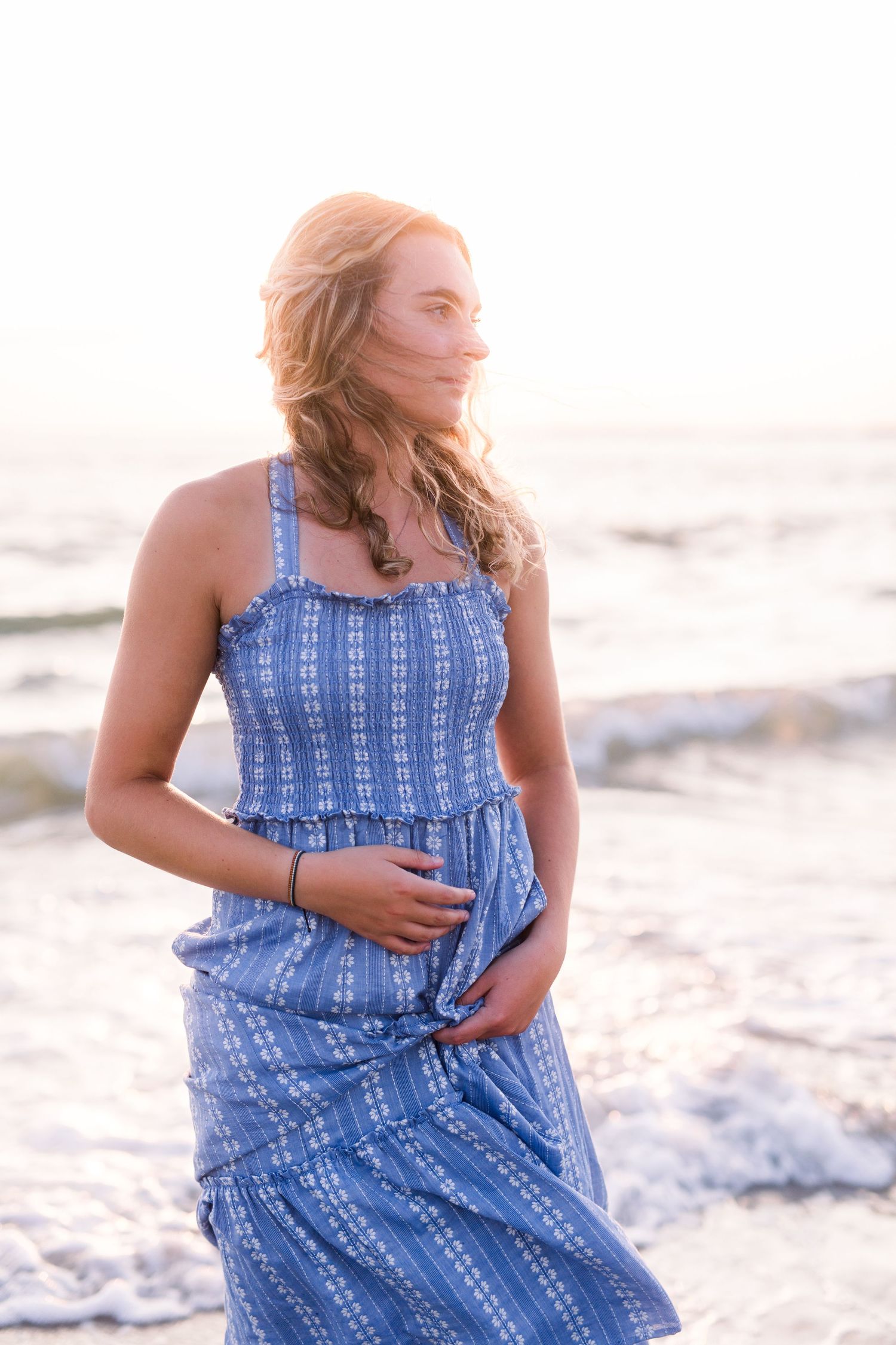 Woman in blue and white patterned sundress standing on beach at sunset with wind blowing through her hair.