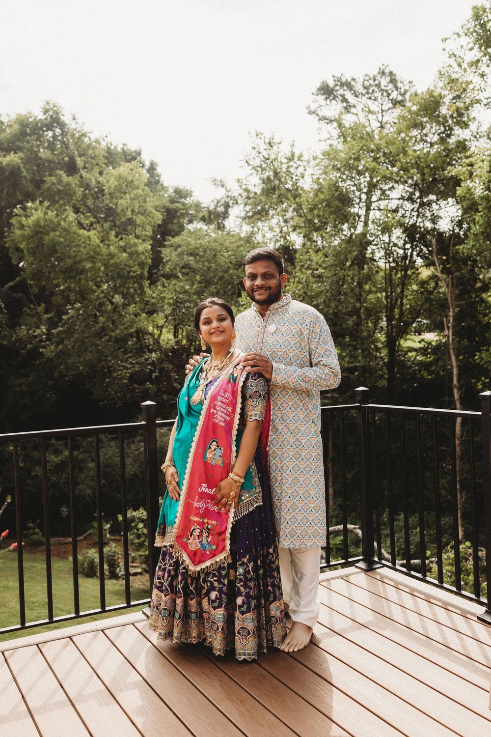 Indian couple in traditional attire posing on a wooden deck surrounded by lush green trees.