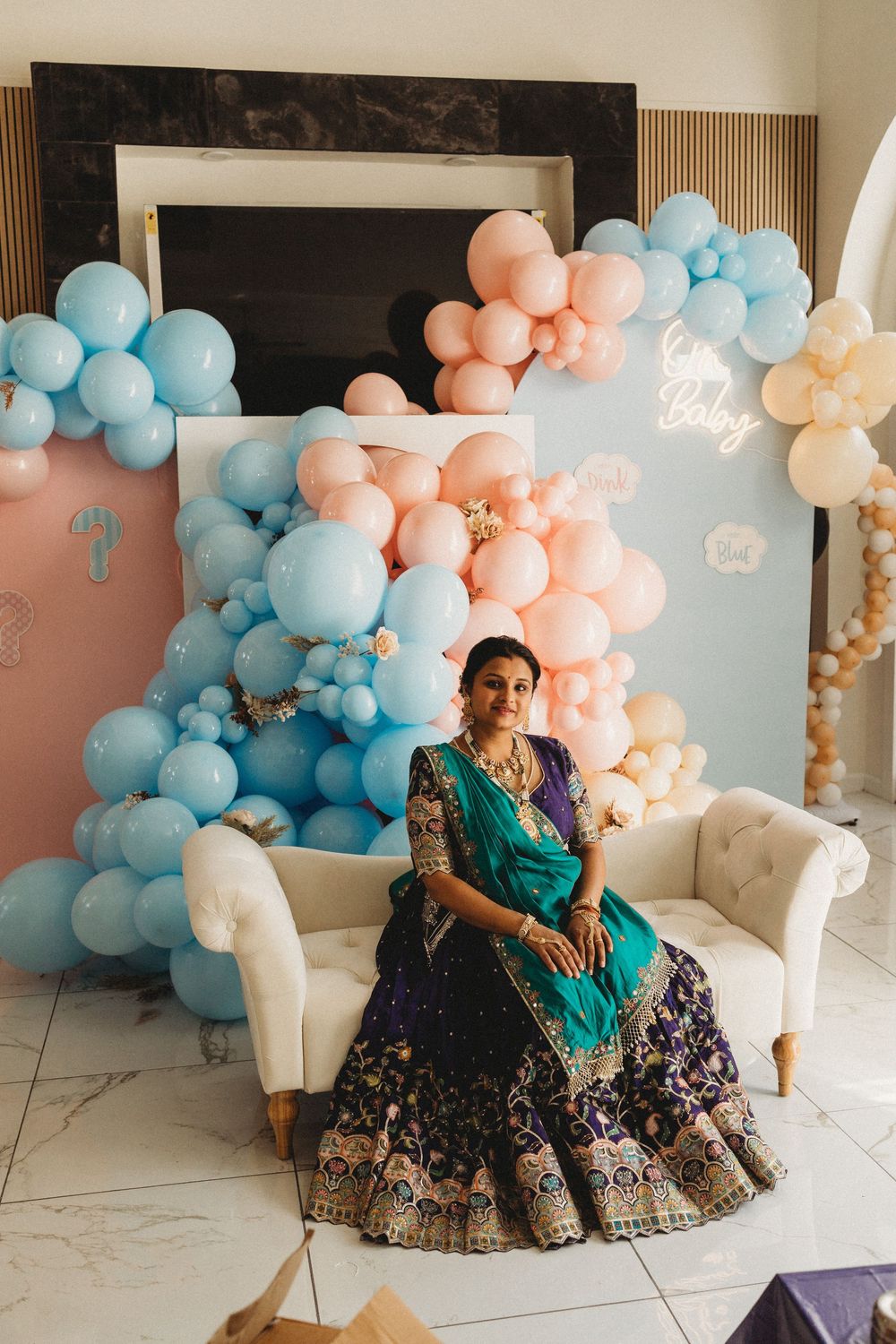 Pregnant Indian woman in purple and teal lehenga sits on white sofa surrounded by pink and blue balloon decorations.