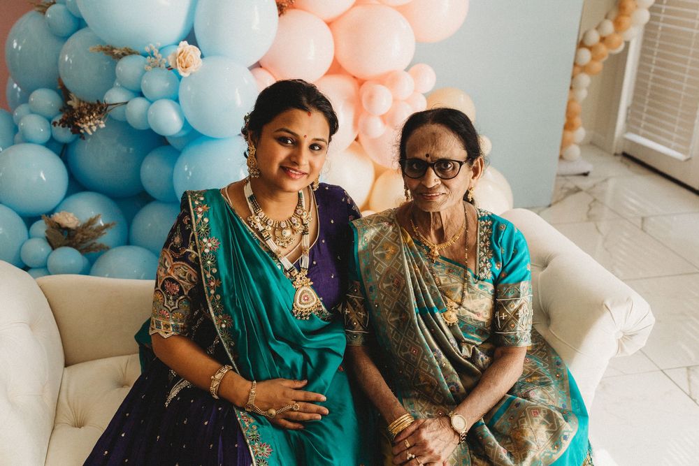 Two women in teal sarees with gold jewelry smile together at a festive celebration with pastel balloons.