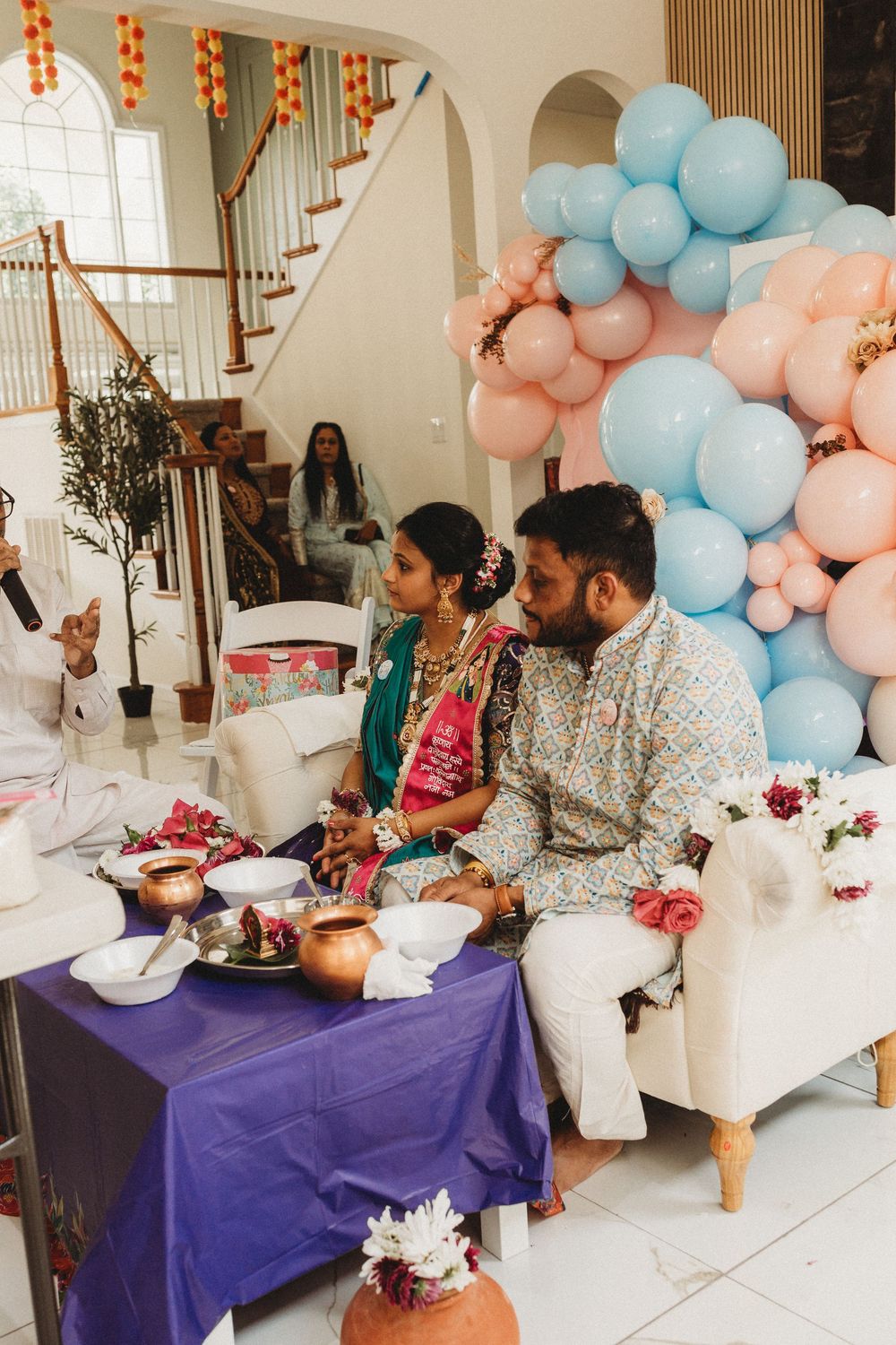 Couple in traditional Indian attire sitting together at a ceremony with colorful balloon arch backdrop.