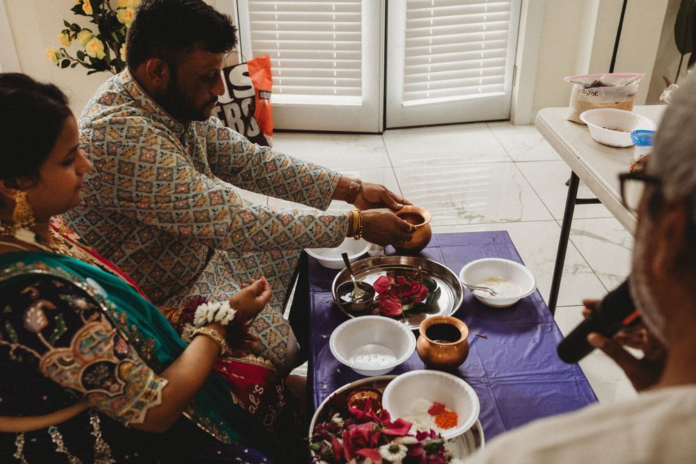 Indian couple performing traditional puja ceremony with flower offerings and ritual bowls on a purple cloth-covered table.