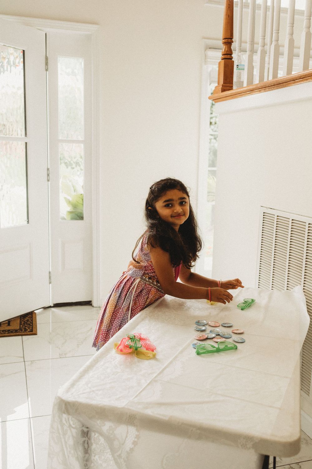 Young girl in pink dress smiling while playing with colorful cookies on a white table indoors.