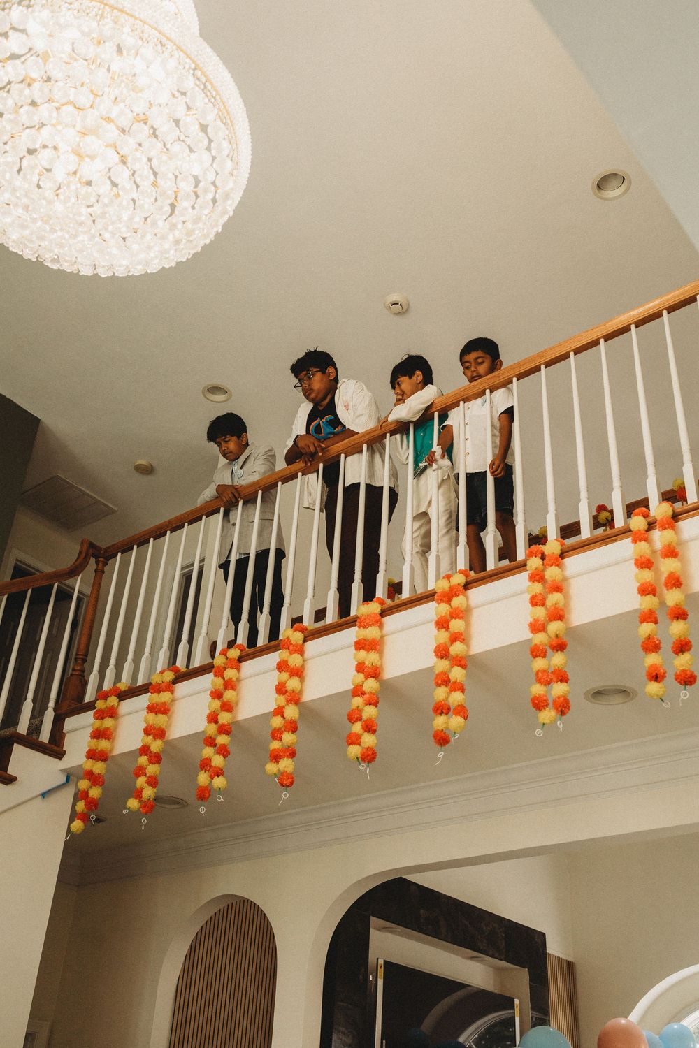 Children look over decorated staircase railing adorned with orange and yellow marigold garlands indoors.