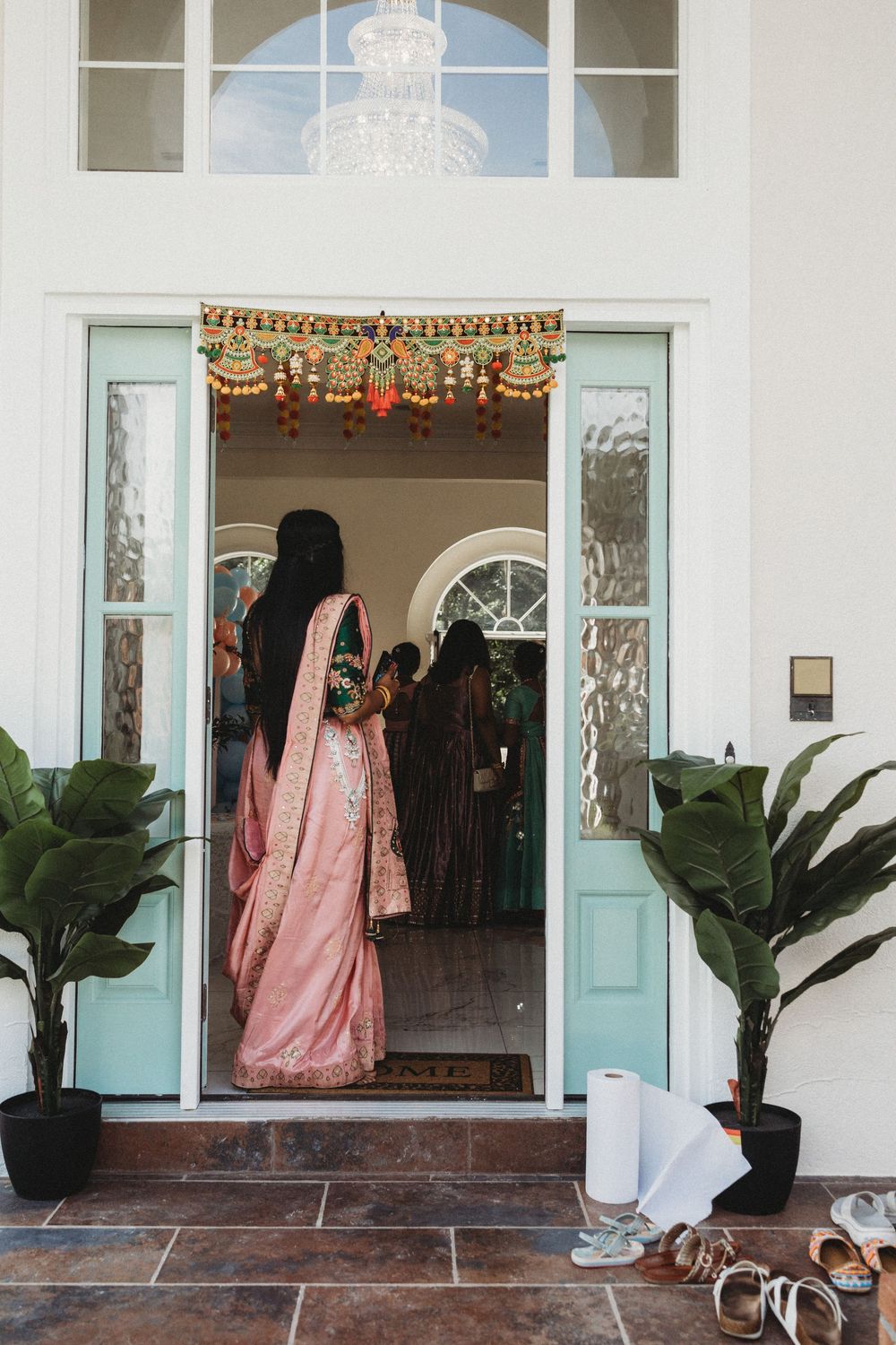 Woman in pink saree stands in doorway adorned with floral toran decoration at traditional Indian wedding ceremony.