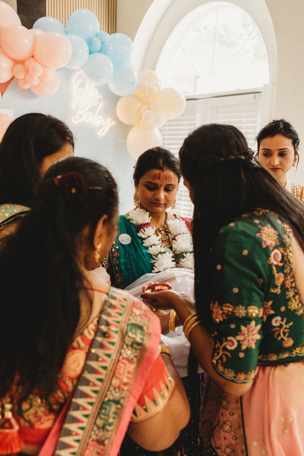 Indian baby shower celebration with women in traditional attire gathered around mom-to-be under pink and blue balloon decor.