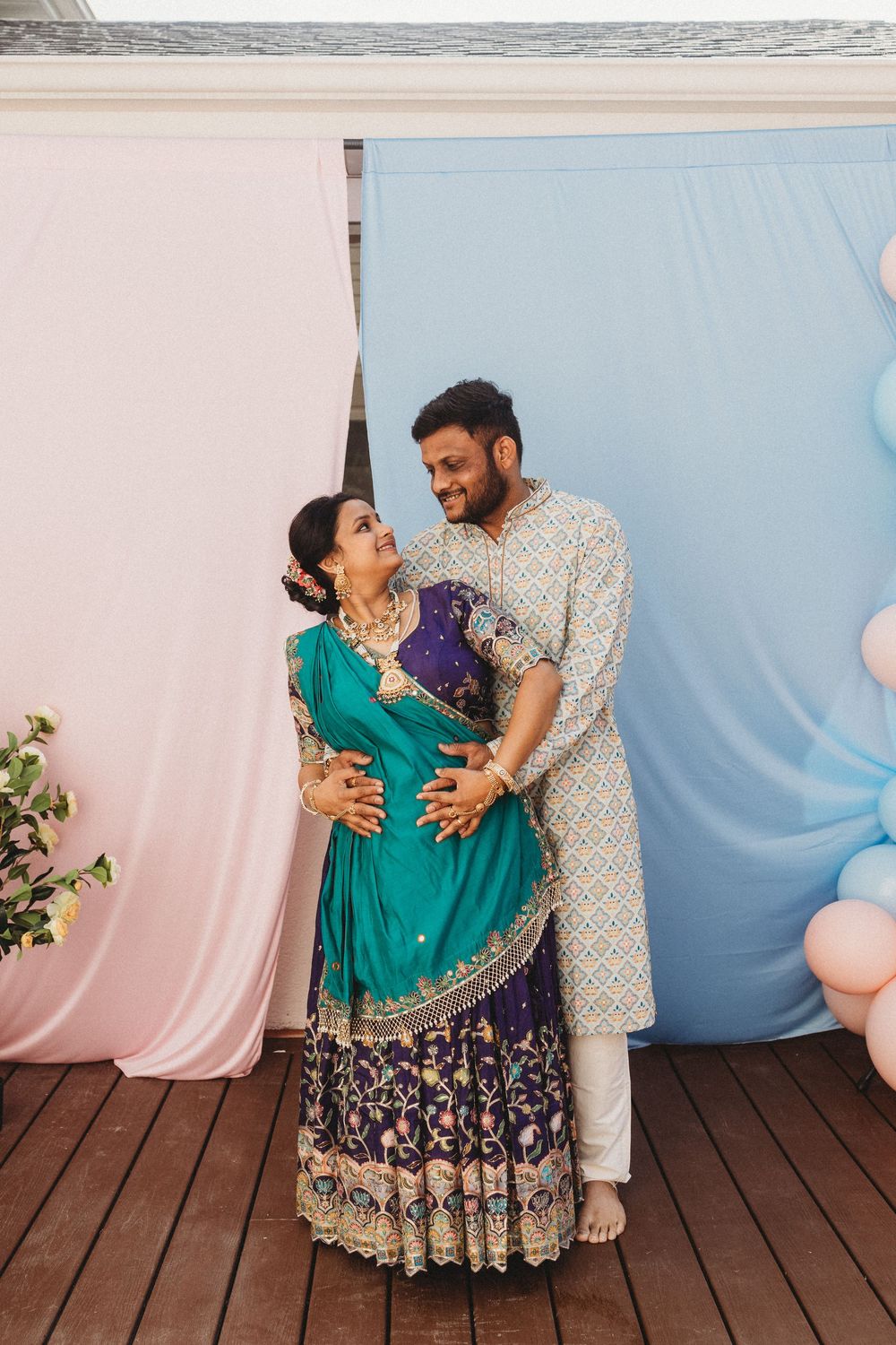 Couple in traditional Indian attire posing together at a gender reveal party with pink and blue backdrop.