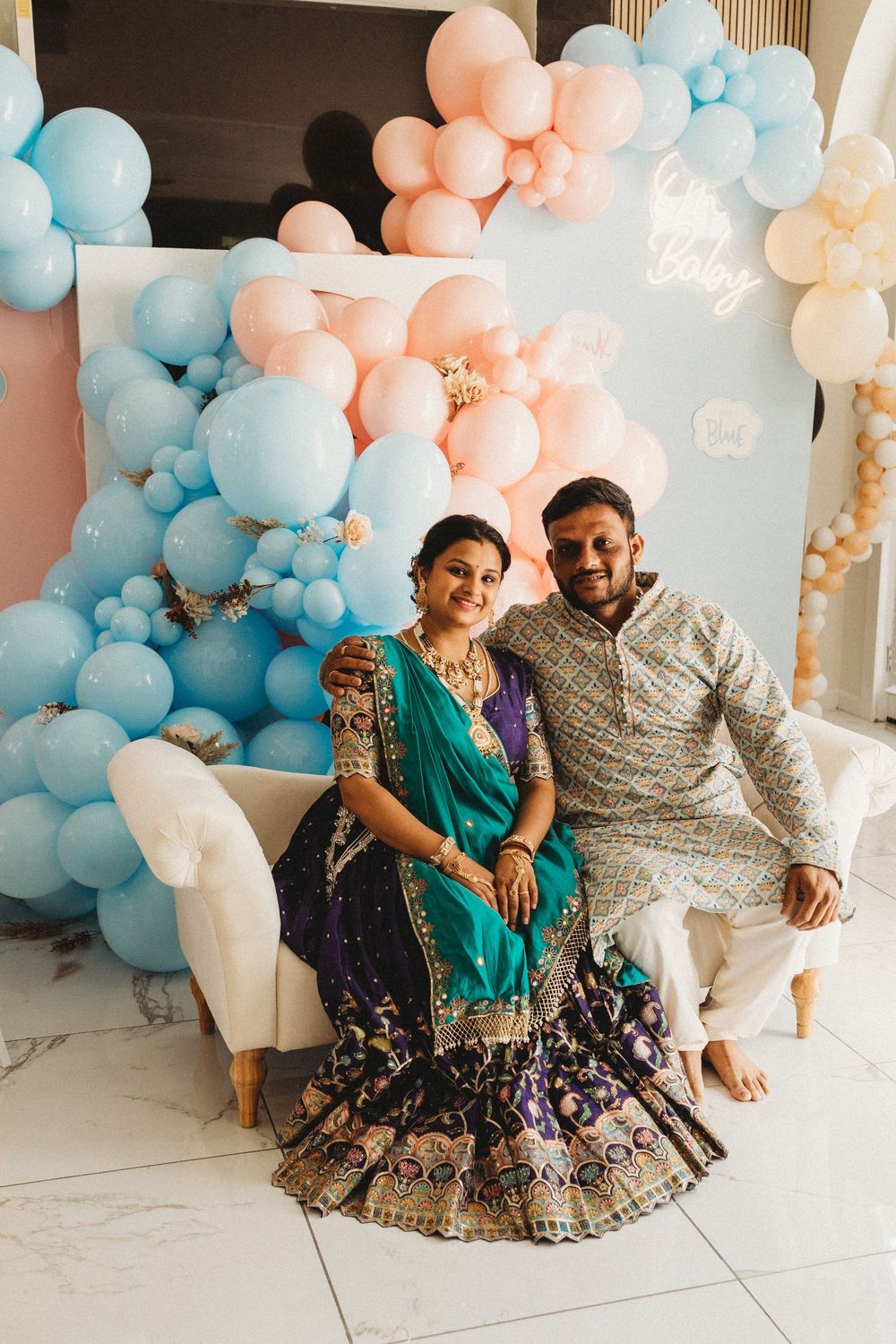 Indian couple in traditional attire seated at a baby shower with blue and pink balloon decorations.