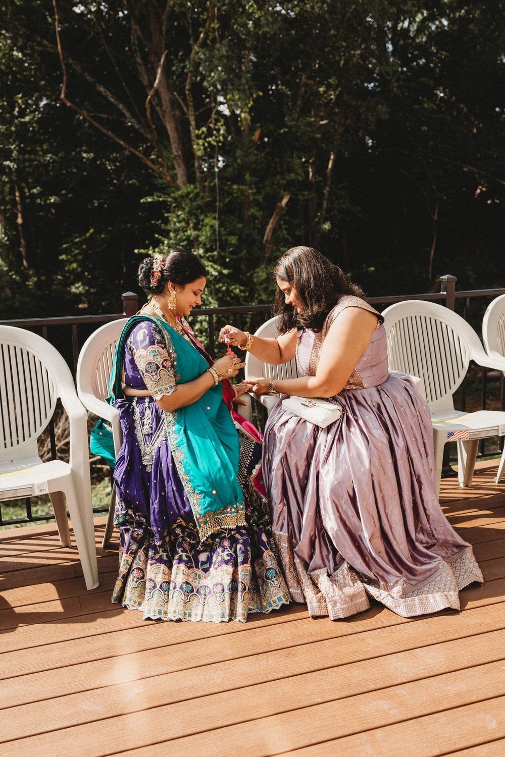 Two women in traditional Indian attire sit on a wooden deck, sharing a moment during an outdoor wedding celebration.