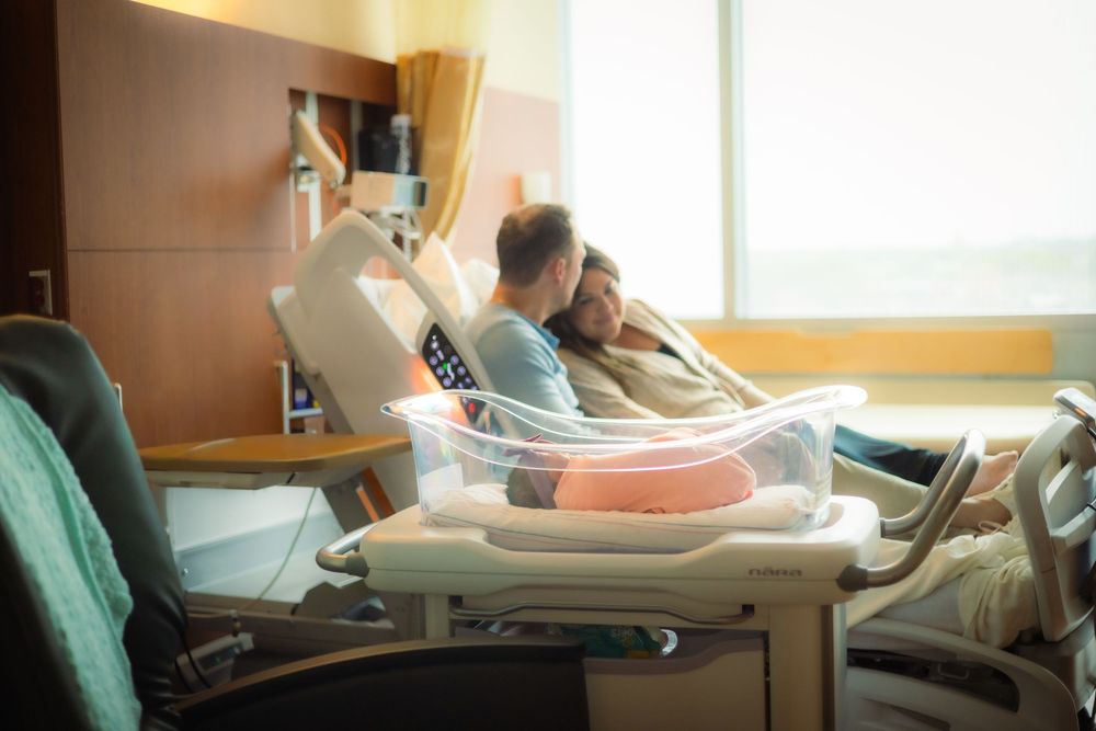 New parents resting together in a hospital room with their newborn baby in a bassinet beside the bed.