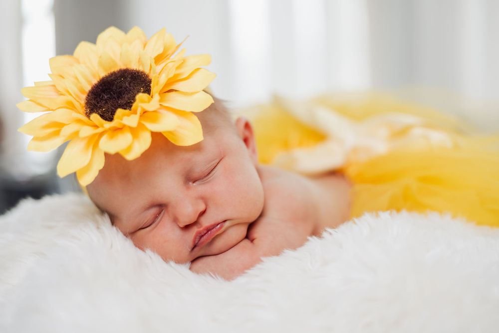 Sleeping newborn baby wearing a yellow sunflower headband on a white fluffy blanket.