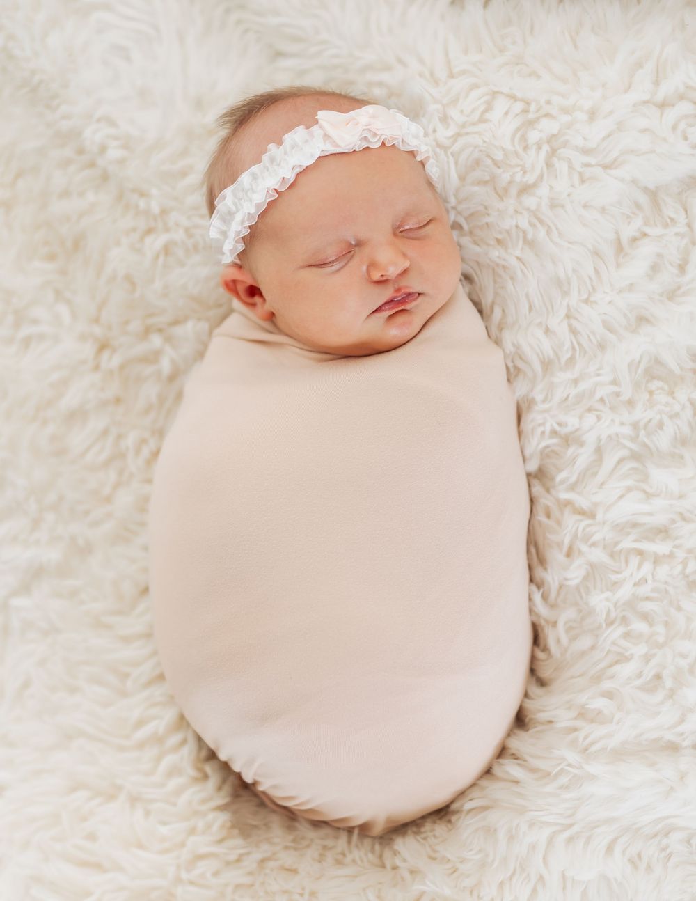 Newborn baby girl swaddled in pink blanket with white floral headband sleeping on fluffy white rug.