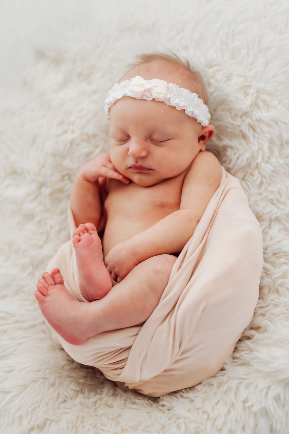 Sleeping newborn baby girl wrapped in beige swaddle with white floral headband on fluffy white blanket.