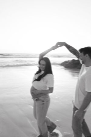 A pregnant woman dances with her partner on a beach in a black and white maternity photo shoot.