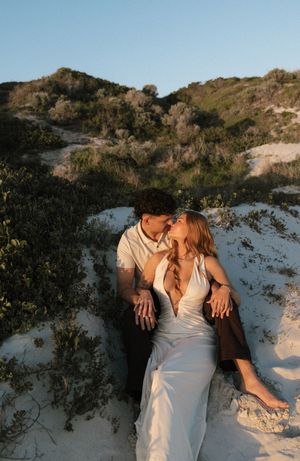 Romantic couple kissing on sandy dunes at golden hour, woman in white dress leaning back.